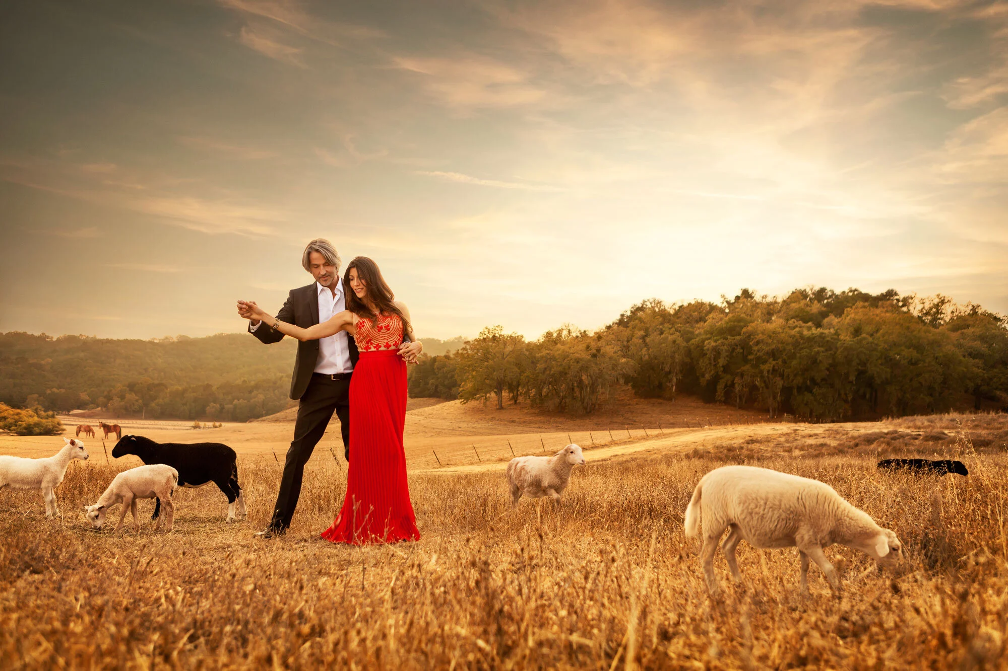 A Latino man in a black suit and woman in a red dress, couple, dancing in a field with sheep and trees in the background at sunset in Paso Robles, CA.