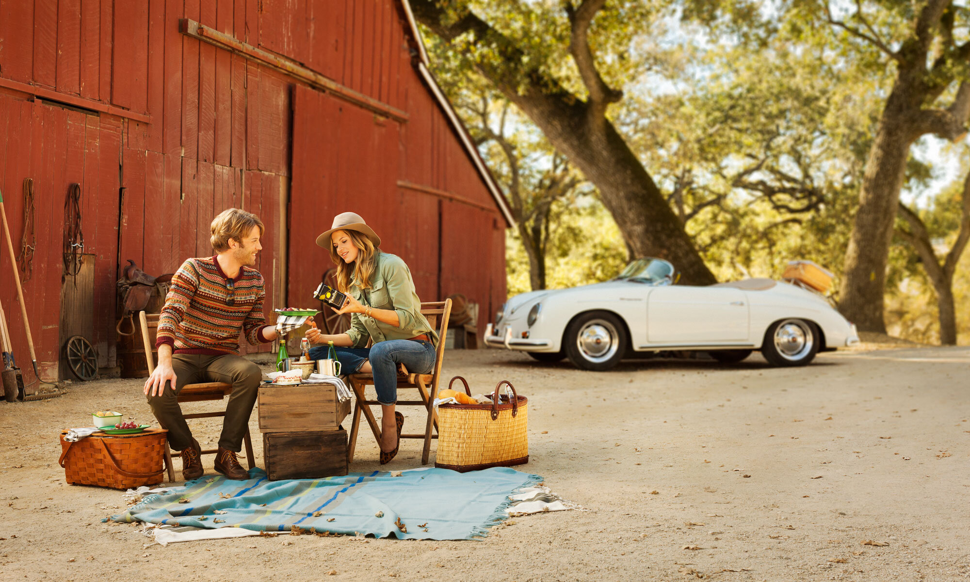 A white couple are having a picnic outside a red barn on a dirt surface with oak trees in the background. The man and woman are sitting  holding a drink and eating. There are baskets, a blanket, and a vintage white convertible car parked nearby at Pa