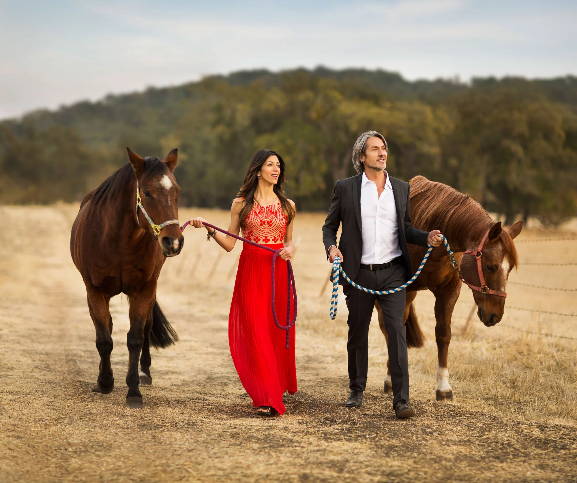 A Latino couple,  walking in a field with two horses. The woman is wearing a red dress and the man is in a black suit with a white shirt, both holding the horses' reins in Paso Robles, CA.