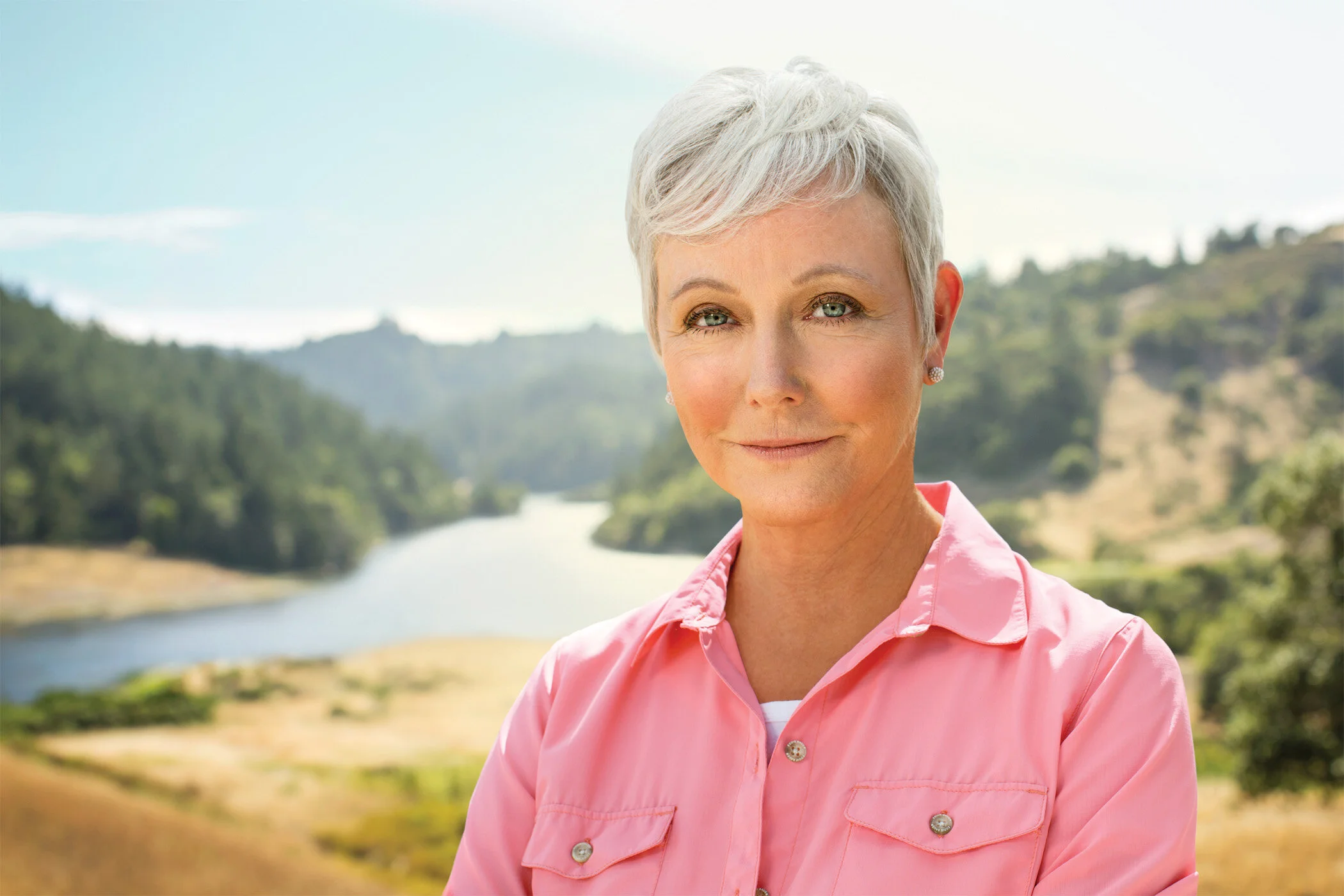 A white mature woman with short gray hair and blue eyes, wearing a pink shirt, standing outdoors with lakes and mountains in the background for Genentech Pharma, Healthcare in San Francisco, CA.
