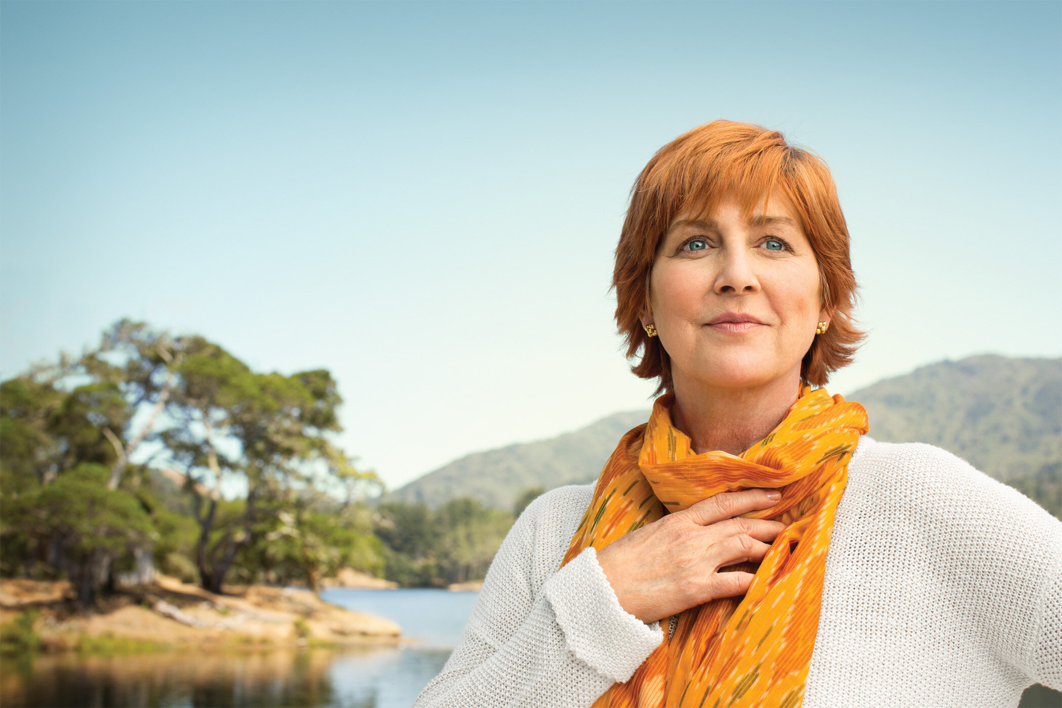 A white mature woman with short red hair and blue eyes, holding her hand over her chest, smiling softly outdoors near a body of water with trees and mountains in the background, wearing white sweater and orange scarf for Genentech Pharma, Healthcare 