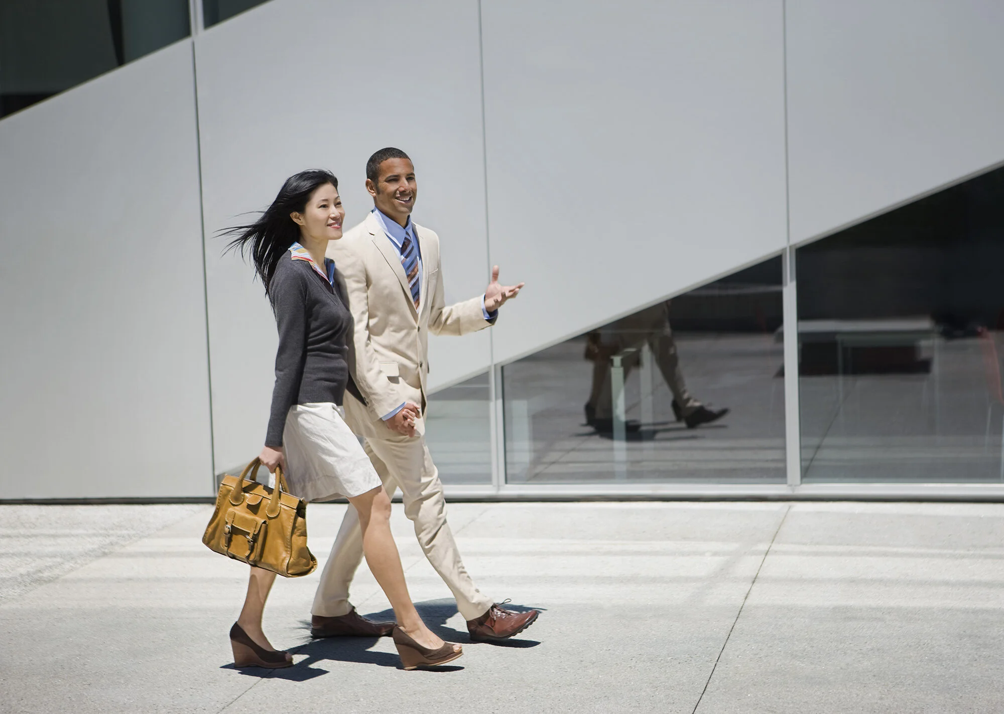Two professionally dressed people, an Asian  woman dressed in a brown shirt and beige skirt, carrying a brown handbag and a Latin man in a tan suit, walking and talking outside a modern building with glass windows at Yerba Buena Park in San Francisco