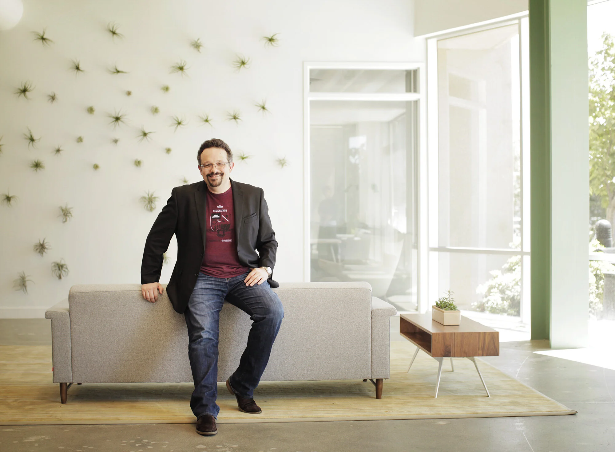 A white, middle aged Phil Libin sitting on a gray sofa in a bright, modern office with white walls, large windows, and a wooden coffee table with a small potted plant at Evernote lobby in Redwood City, CA.