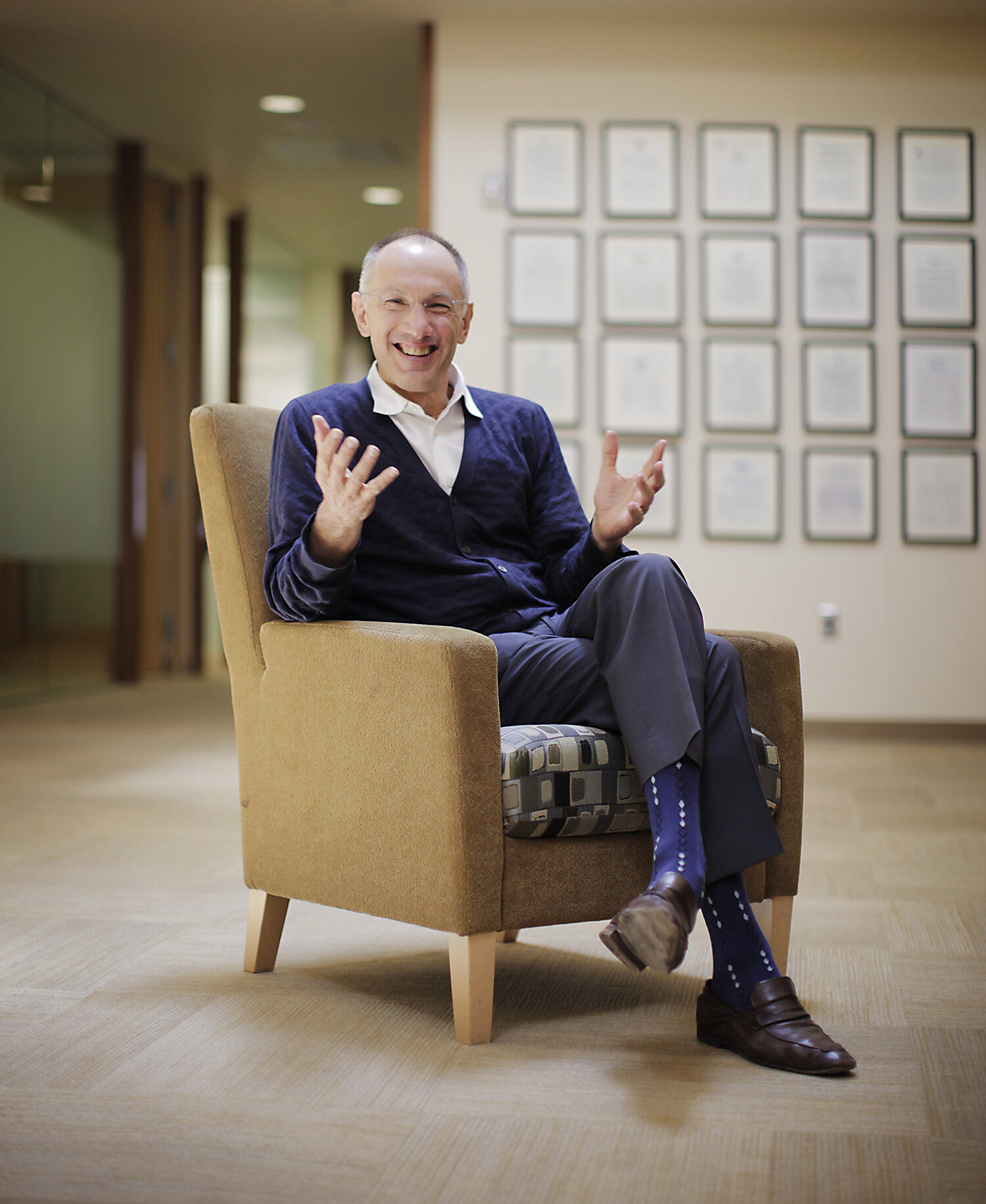 A white, middle aged Welsh business man, investor and venture capitalist, Michael Moritz, smiling and gesturing while seated in a beige armchair at office lobby with framed certificates on the wall at Sequoia Capital in Menlo Park,  CA.