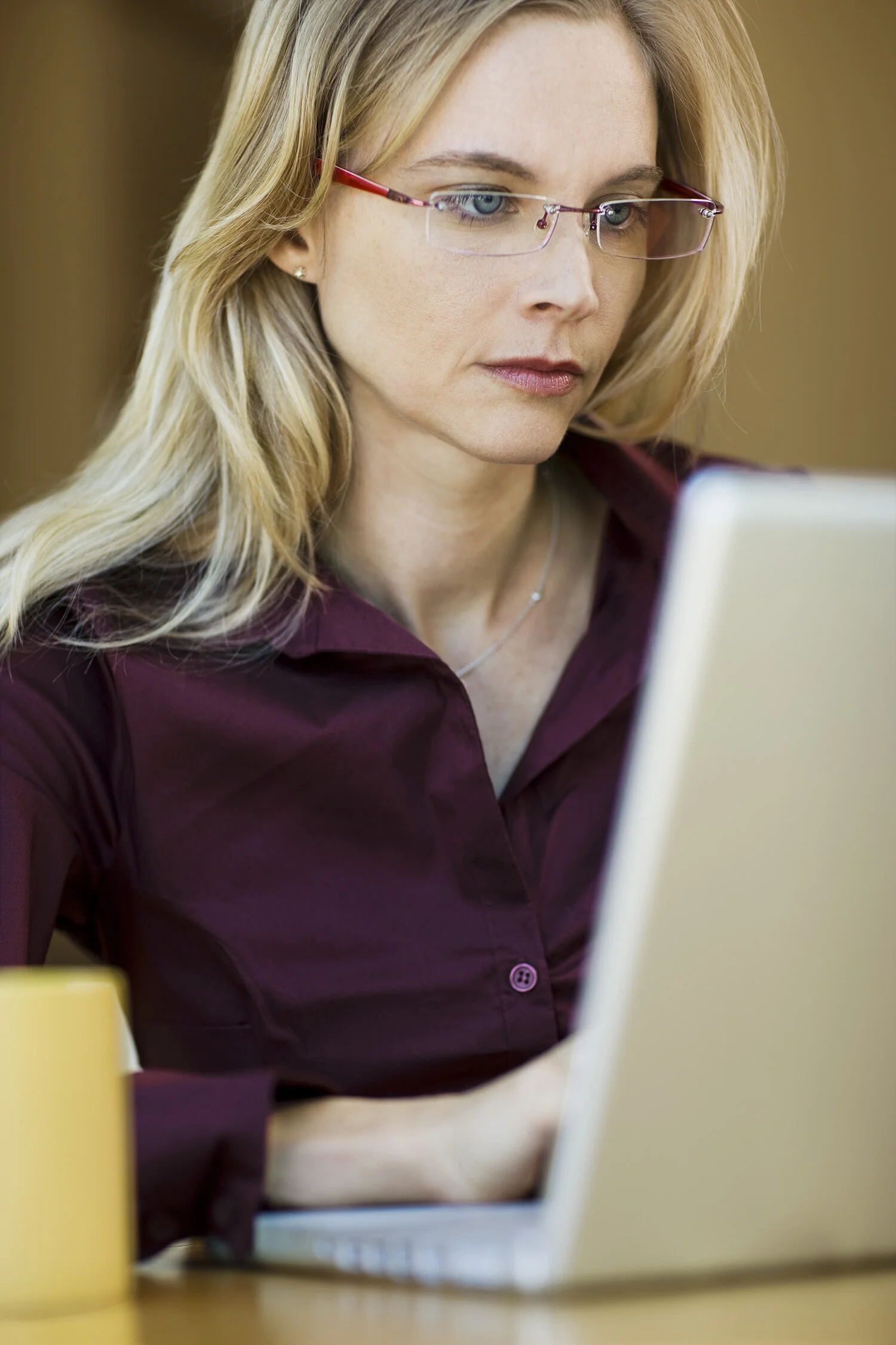 A white woman with long, blonde hair and glasses working on a laptop, wearing a burgundy shirt, with a yellow mug on the table in San Francisco, CA.