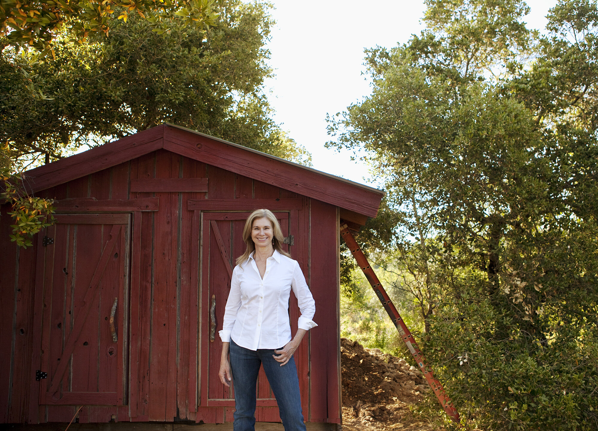 A white farmer woman smiling in a white shirt and jeans standing in front of a small red wooden shed with trees in the background in Sonoma, CA.