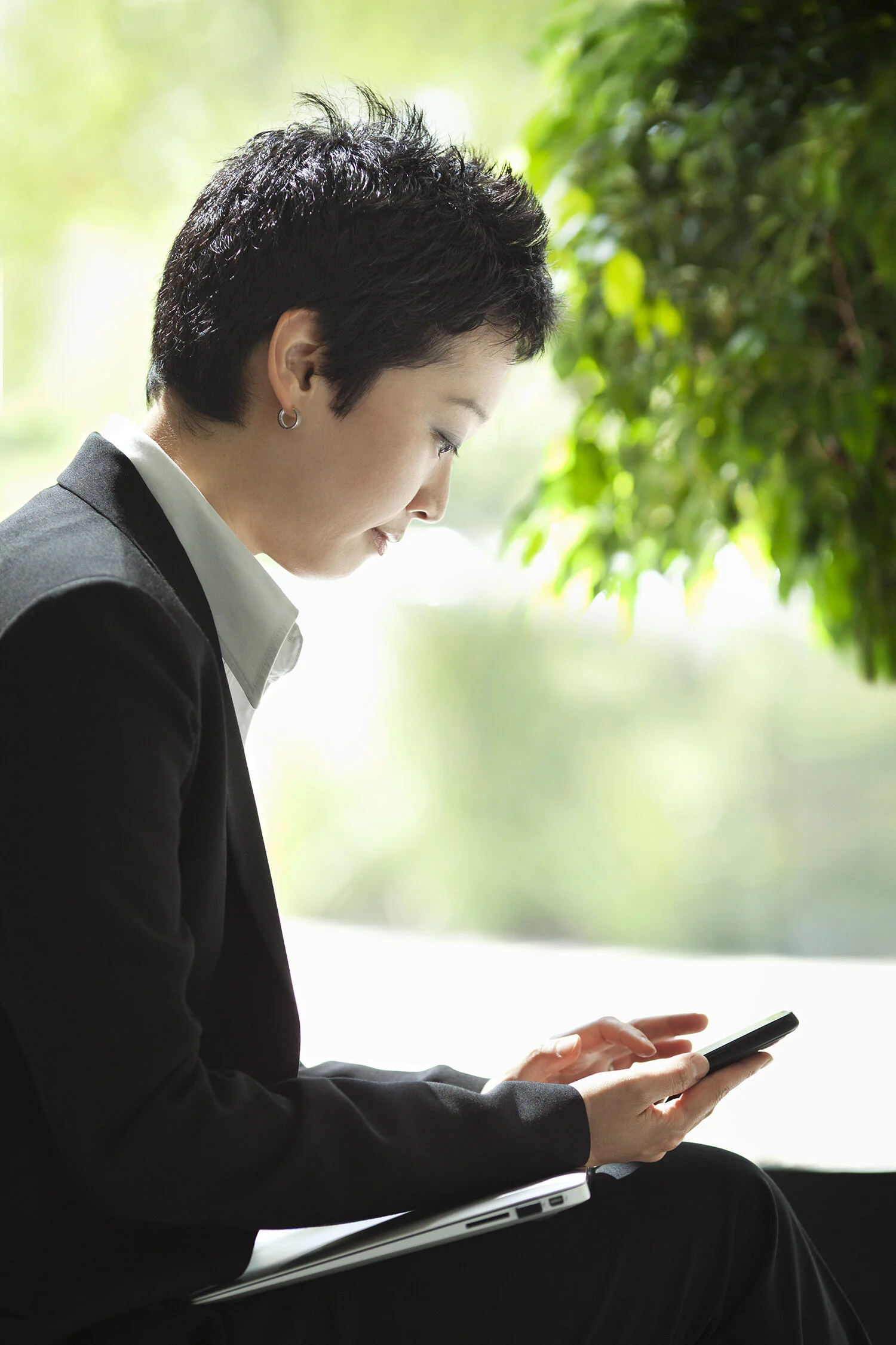 An Asian woman in black business attire, sitting and looking at her smartphone, with a closed laptop on her lap, near a window with green foliage outside at Cisco in San Jose, CA.