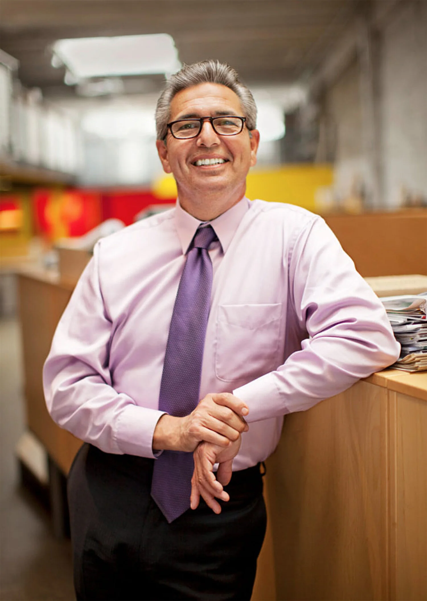 A smiling middle-aged man with glasses, wearing a light pink shirt and purple tie, leaning on a wooden counter in an architectural  office environment in San Francisco, CA.