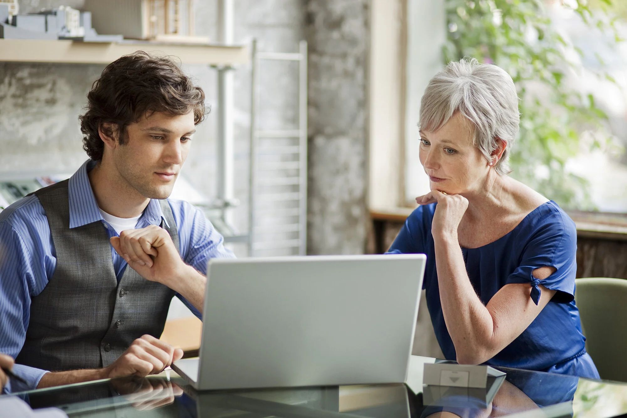 A young man with brown, short hair in a blue shirt and a grey vest, and mature woman in a blue dress and short, grey hair, sitting at a table in a bright architectural room, looking at a laptop screen with thoughtful expressions in San Francisco, CA.