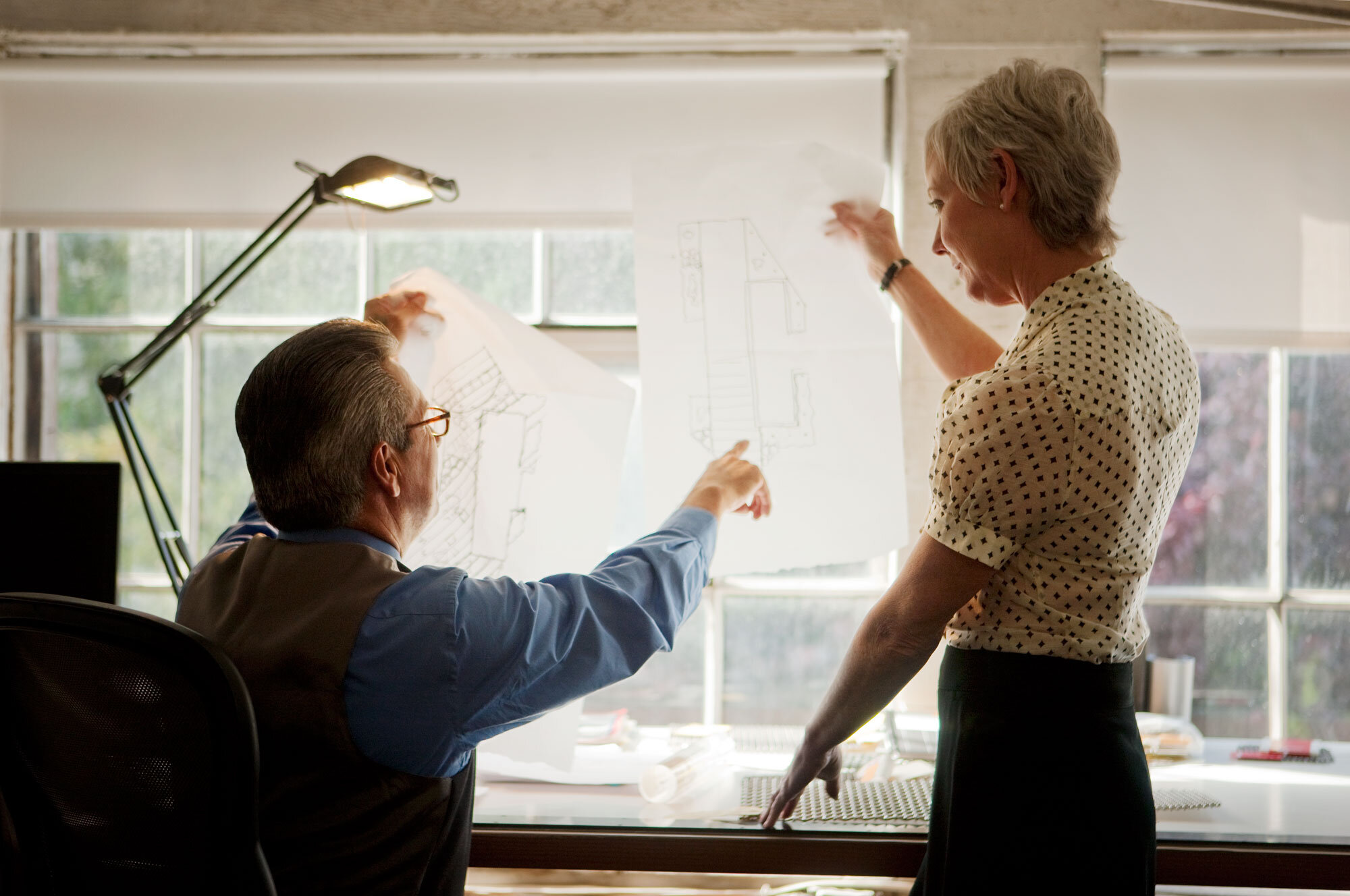 Two white, middle aged professionals in an office with a lamp on the table discussing construction plans with blueprints on a windowed wall in architectural office in San Francisco, CA.