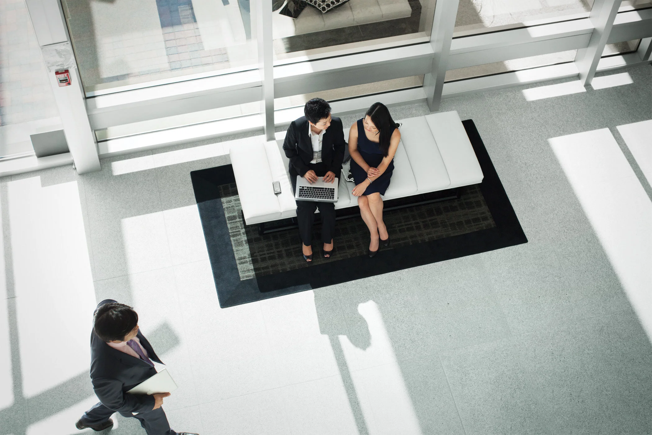 Bird's eye view of two Two Asian women in business attire sitting on a white bench in a modern office lobby, talking, looking at an Apple laptop with a man walking past holding a folder at Cisco, in San Jose, CA.
