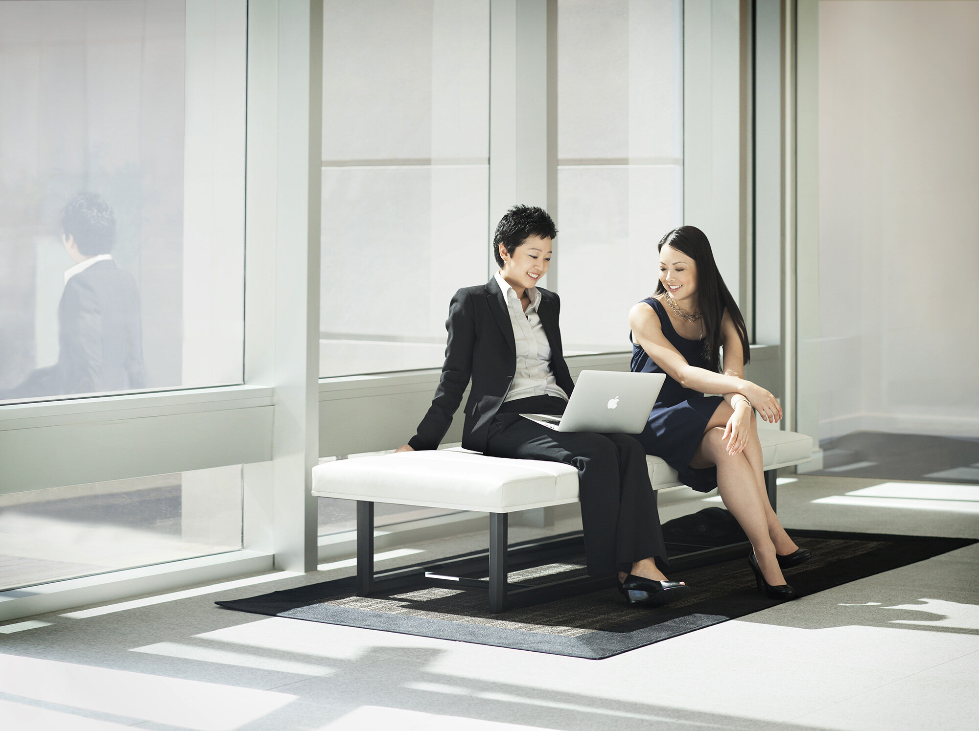 Two Asian women in business attire sitting on a white bench looking at an Apple laptop against large windows in a modern office lobby smiling at Cisco, San Jose, CA.