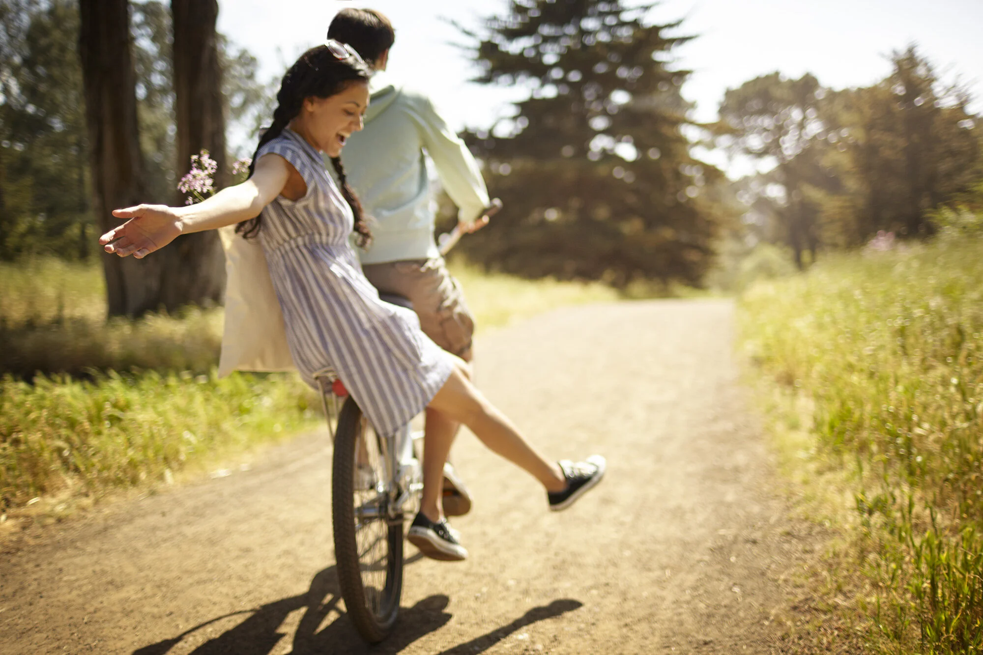 A young Latina woman in a summer dress sitting on the back of a bicycle with a young man biking on a dirt trail in a park, with sunlight filtering through the trees in San Francisco, CA.