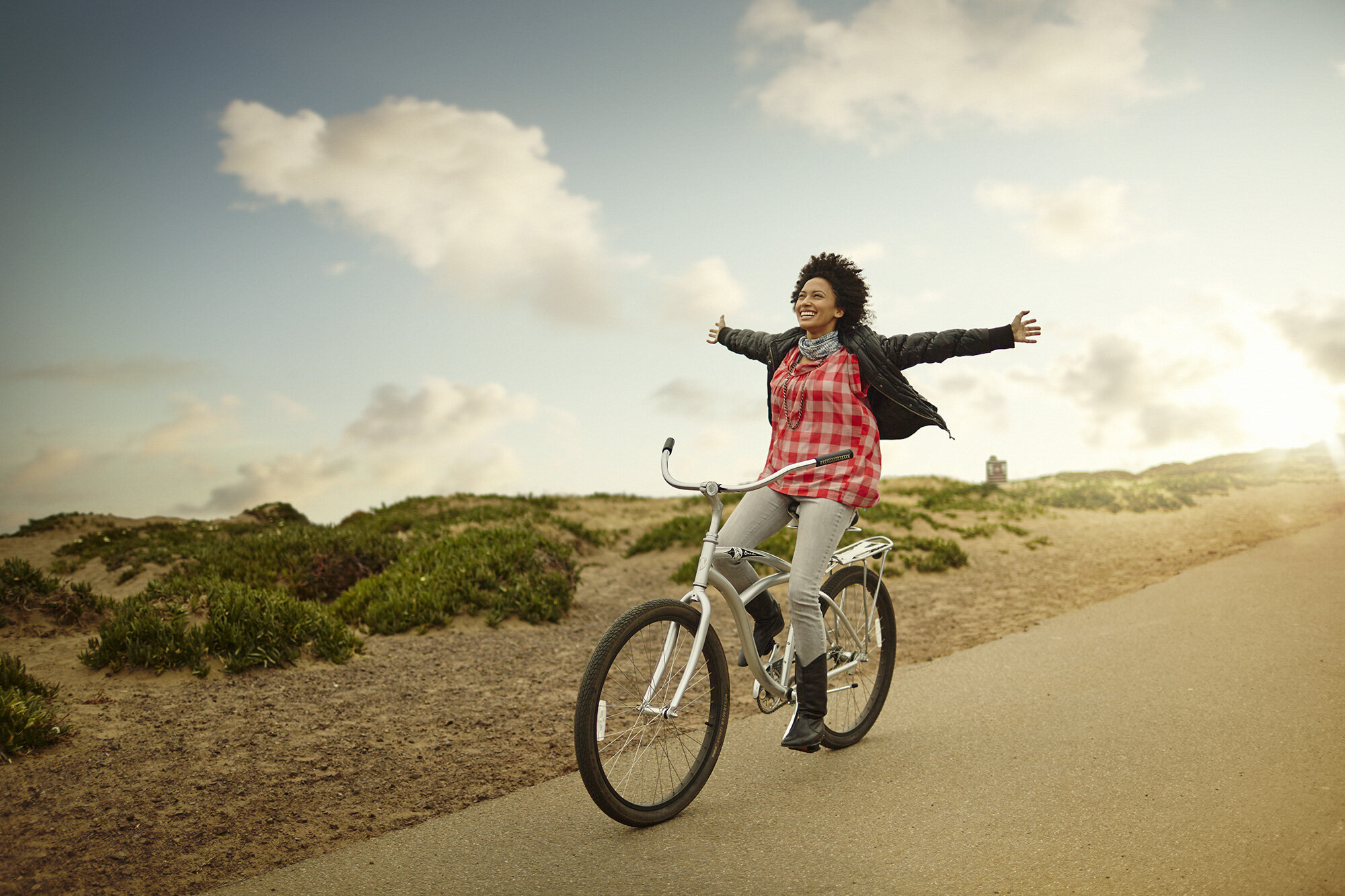 A young Black woman woman riding a bicycle with arms outstretched in a checkered red and white shirt on a path in a natural landscape with bushes and a partly cloudy sky in San Francisco, CA.