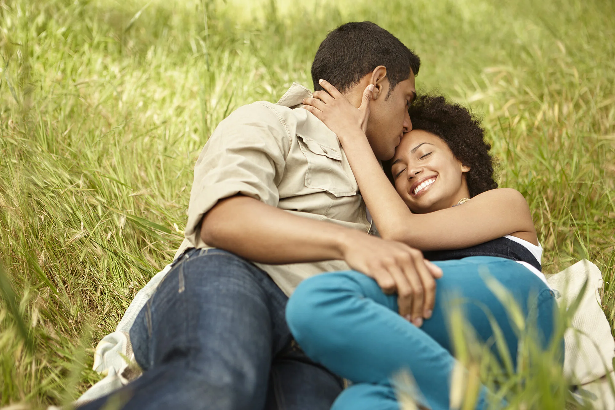 A couple, Black woman and Latino man sitting in a grassy field, embracing and smiling, with the man kissing the woman's forehead in San Francisco, CA.