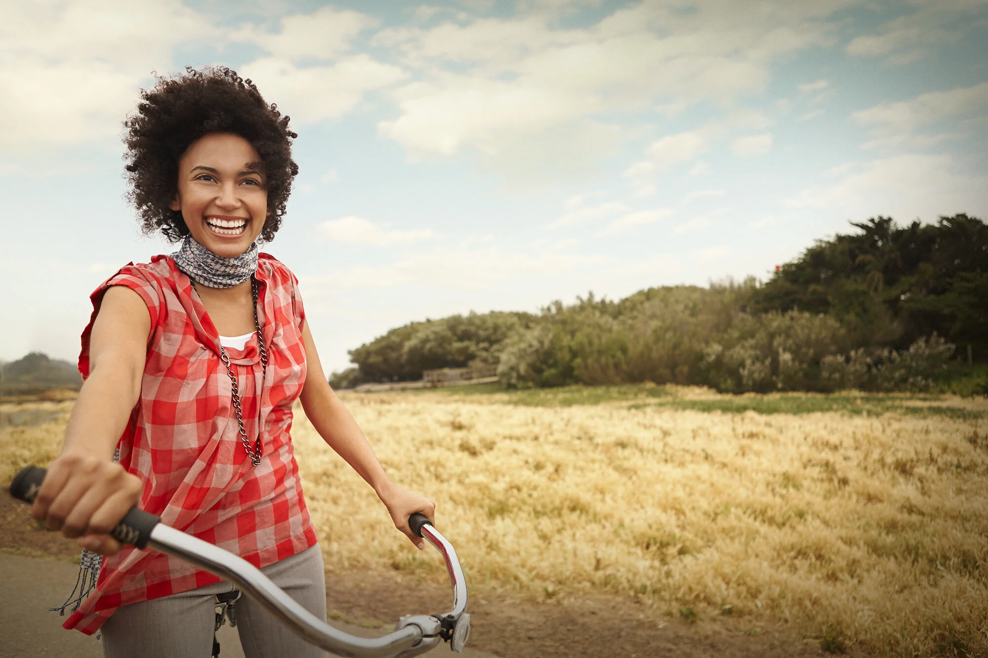 A young, Black woman riding a bicycle outdoors, smiling and wearing a red and white checkered shirt with a necklace and scarf, in a rural setting with fields and trees under a partly cloudy sky in San Francisco, CA.