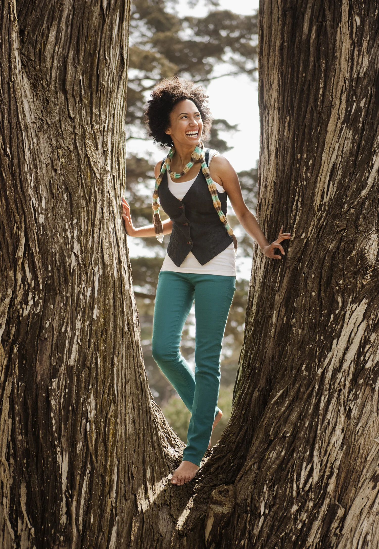 A young Black woman with curly hair smiling in green pants and black vest while standing between two large tree trunks in an outdoor setting in San Francisco, CA.
