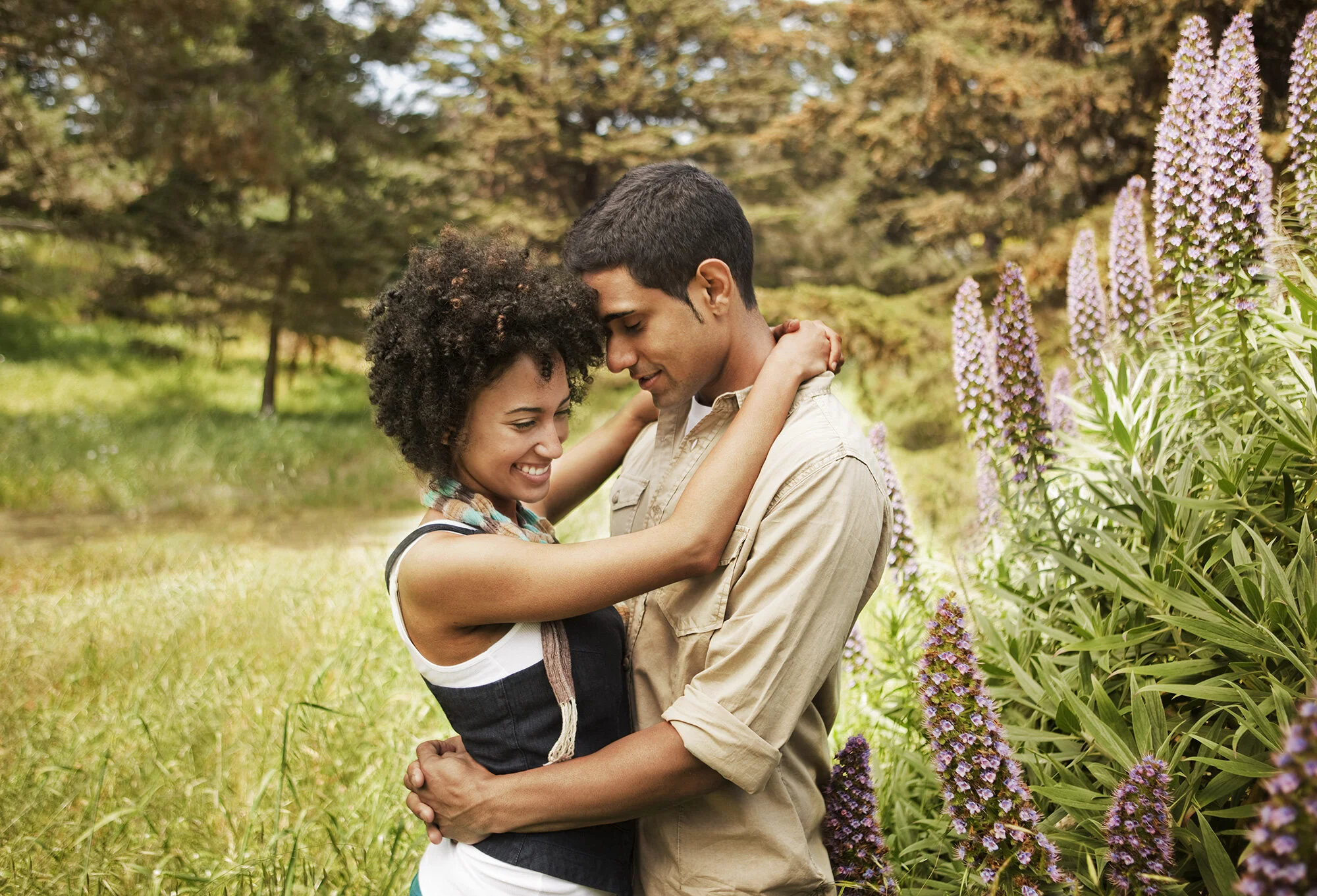 A smiling couple, Black woman and Latino man hugging in a green park surrounded by tall purple flowers in San Francisco, CA.