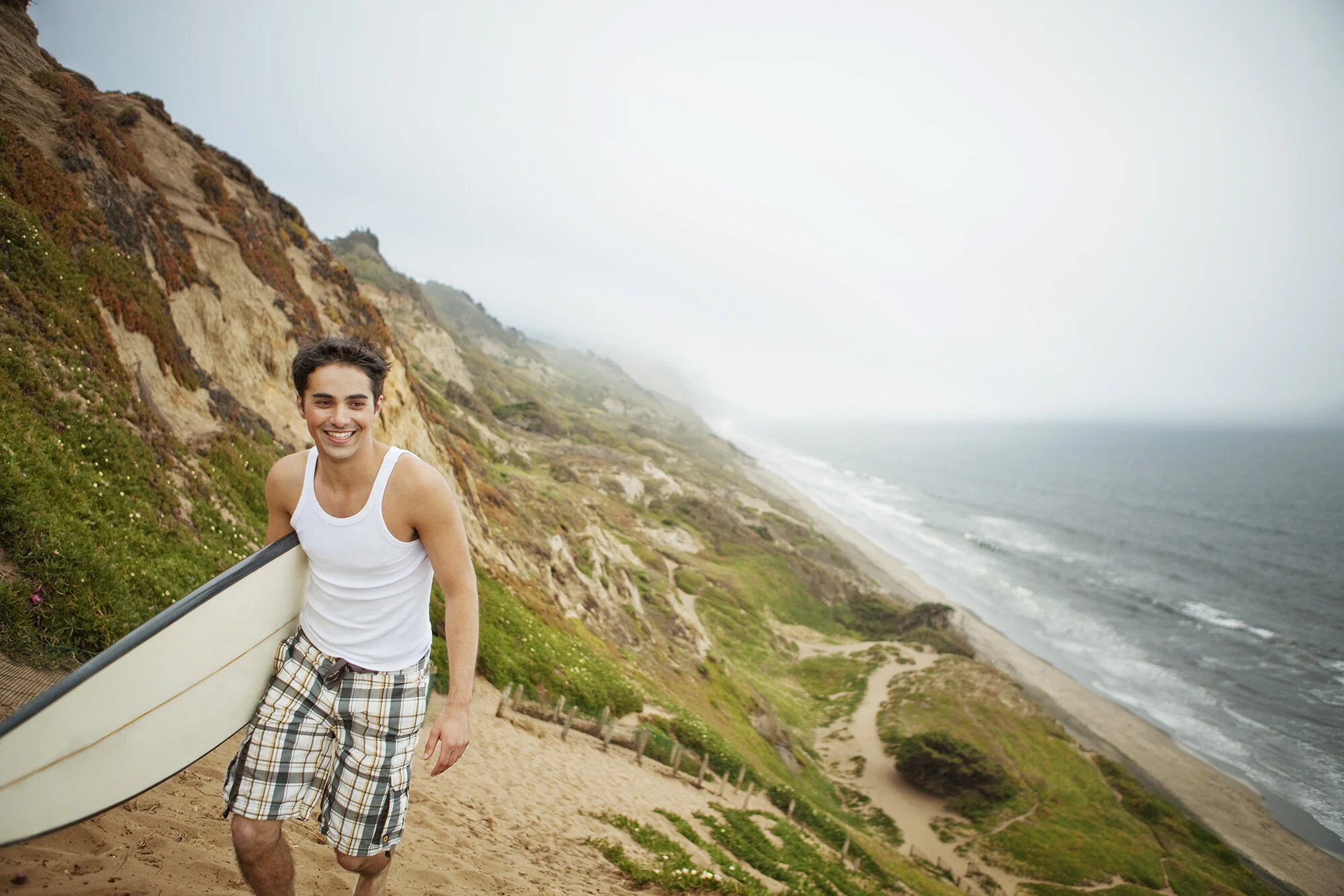 Young white man carrying a surfboard smiling on a sandy trail near a grassy cliffside by the ocean on a foggy day at Fort Funston in San Francisco, CA.