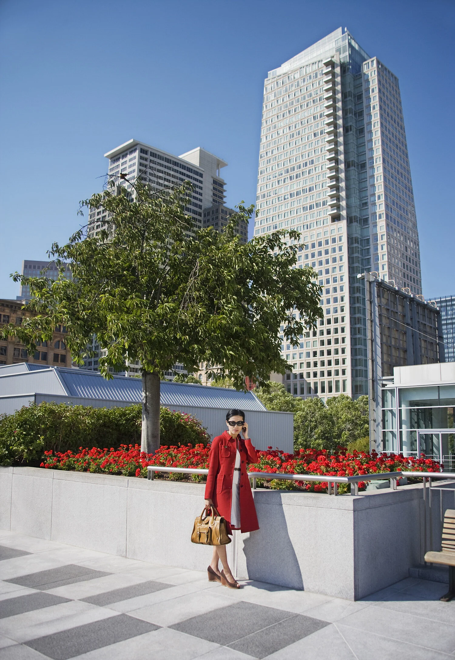 An Asian woman in a red coat and sunglasses is standing outdoors in an urban park with a brown bag and high heels, talking on her cellphone, with tall modern office buildings in the background at Yerba Buena Park in San Francisco, CA.