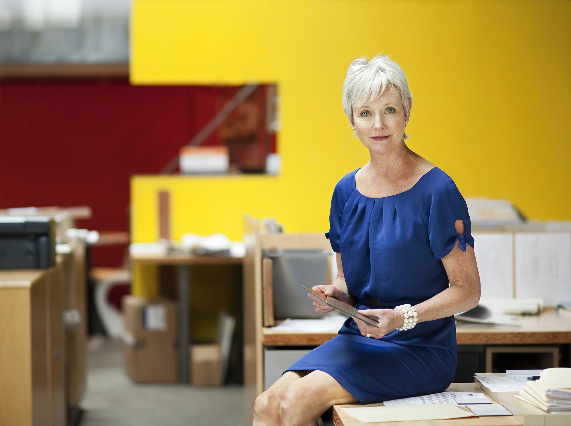 A middle aged woman with short gray hair in a blue dress sitting on a desk in an architectural office with a yellow wall, holding a tablet and looking at the camera in San Francisco, CA.