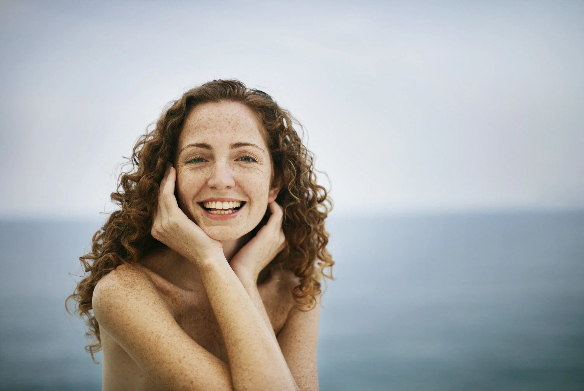 A young, white, Brazilian woman with curly red hair and freckles smiling with her hands on her cheeks, outdoors by the ocean at Ipanema beach in Rio de Janeiro, Brazil.