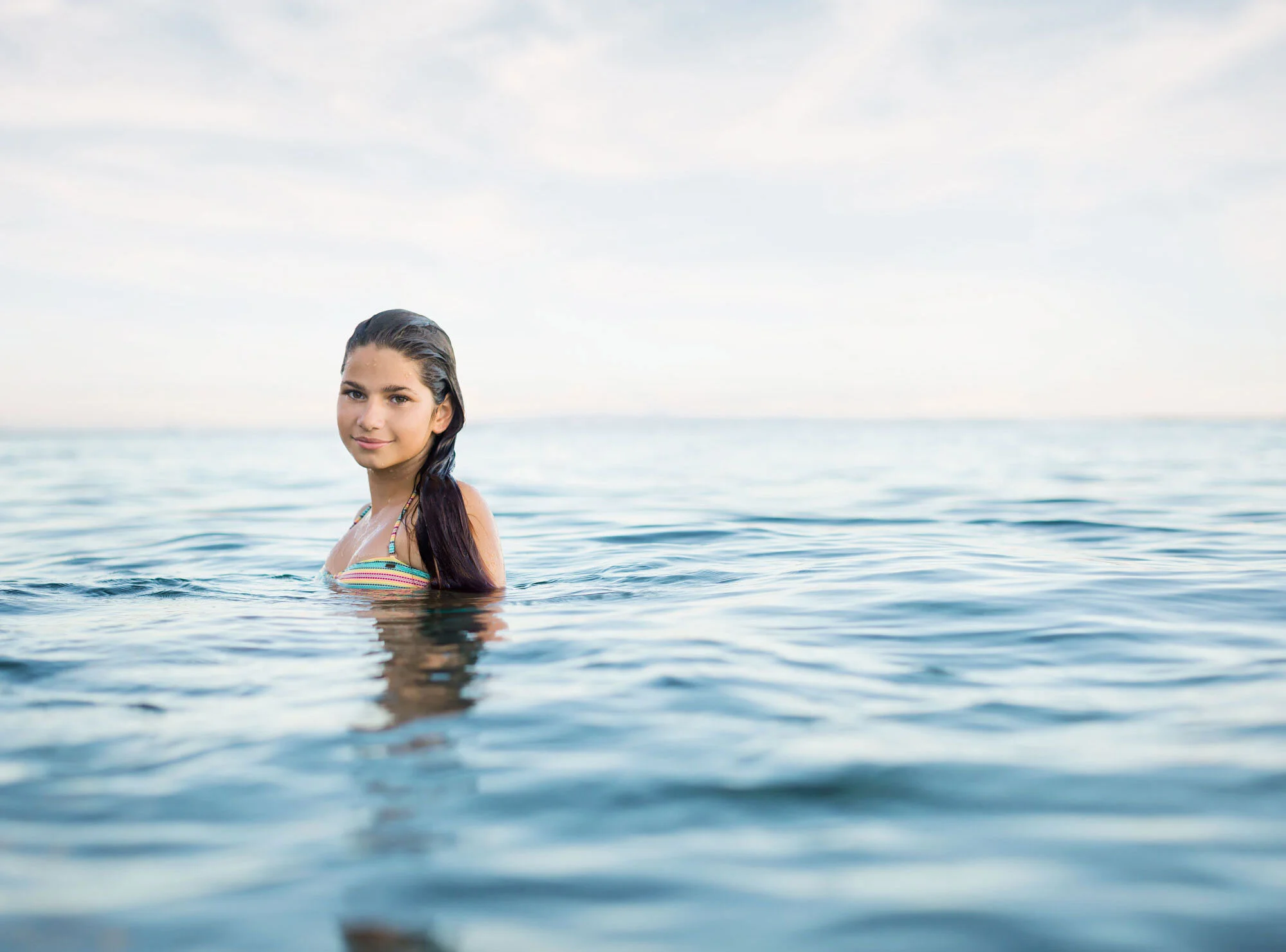 A young white woman with long dark hair is swimming in the ocean during the daytime, wearing a striped swimsuit with beads on the straps, with a calm water surface and a partly cloudy sky in the background in Maui, Hawai'i.