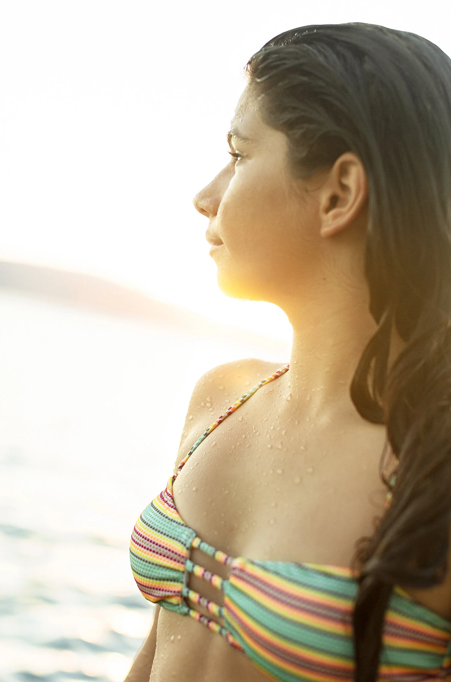 Side profile of a young girl with wet hair, wearing a colorful striped bikini top, standing by the water at sunset in Maui, Hawai'i.