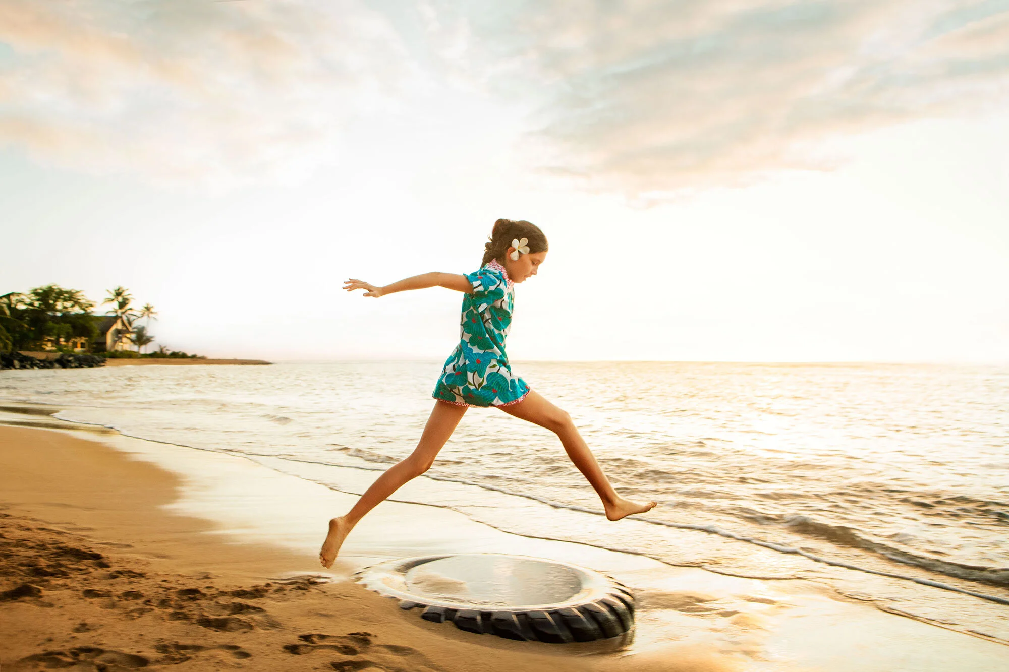 A white girl in a colorful dress with a flower in her hair jumping on a tire on the beach at sunset at a beach in Maui, Hawai'i.