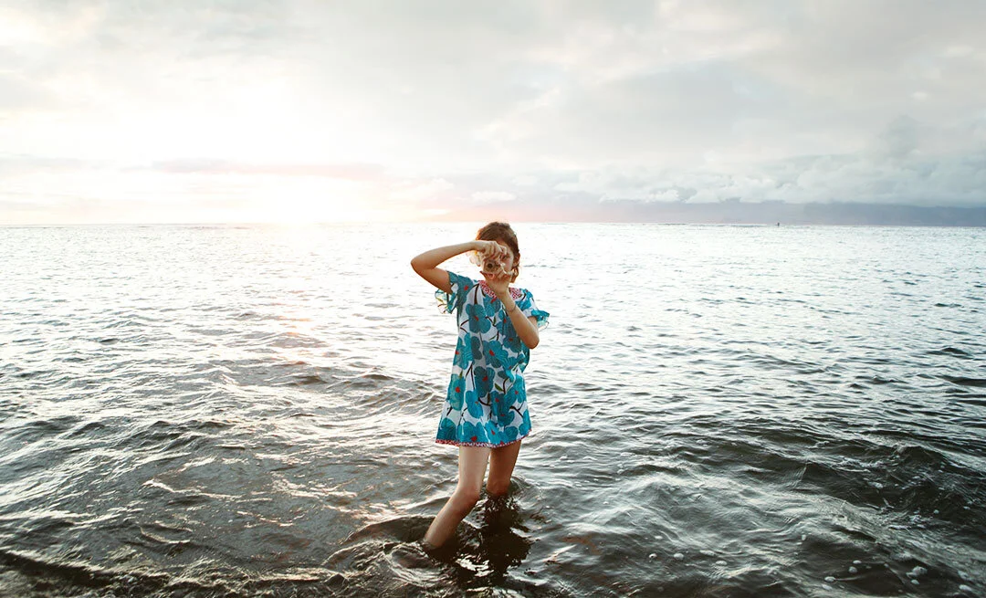 A white girl standing in the ocean wearing a blue floral dress, holding a seashell up to her eye, with a sunset in the background in Maui, Hawai'i.