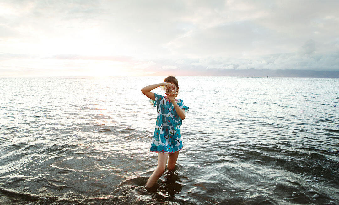 A white girl standing in the ocean wearing a blue floral dress, holding a camera up to her eye, with a sunset in the background in Maui, Hawai'i.