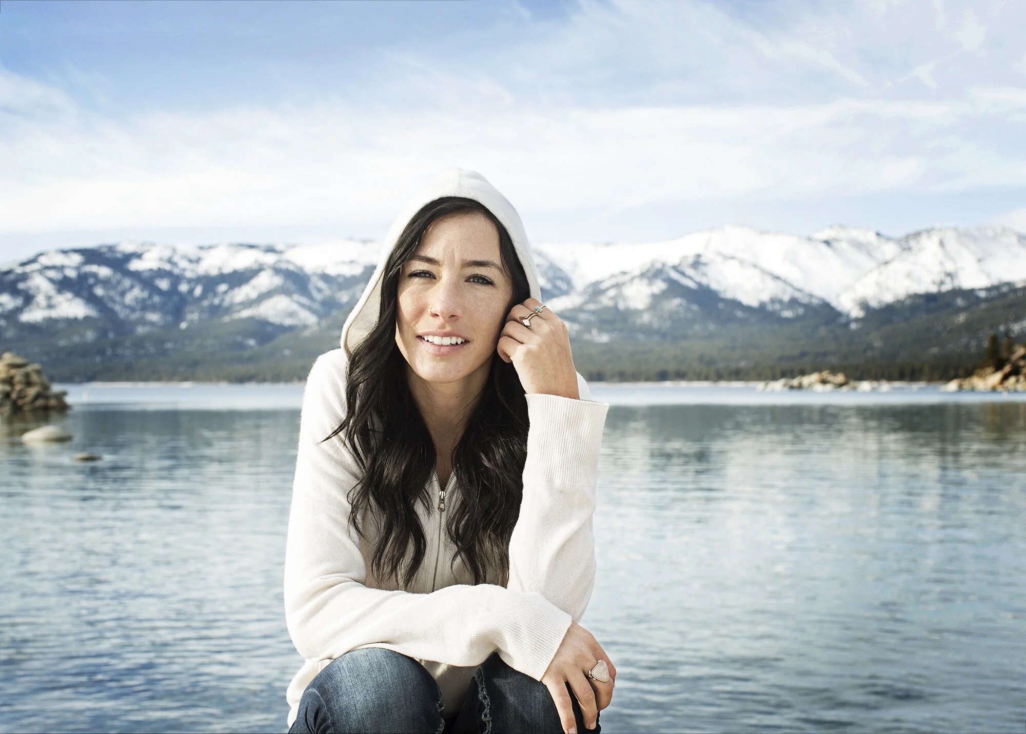A white woman with long black hair and blue eyes, wearing a a white hoodie and blue jeans smiling with a mountain landscape and lake in the background at Sand Harbor in Lake Tahoe, CA.