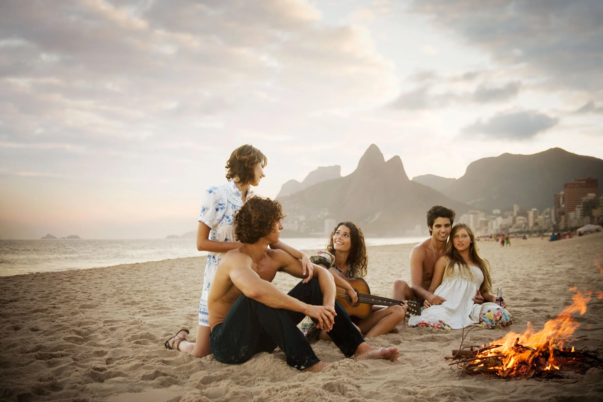 Six young adults sitting on Ipanema beach during sunset, with some playing guitar and others chatting, near a small campfire, with mountains and city buildings in the background in Rio de Janeiro, Brazil.
