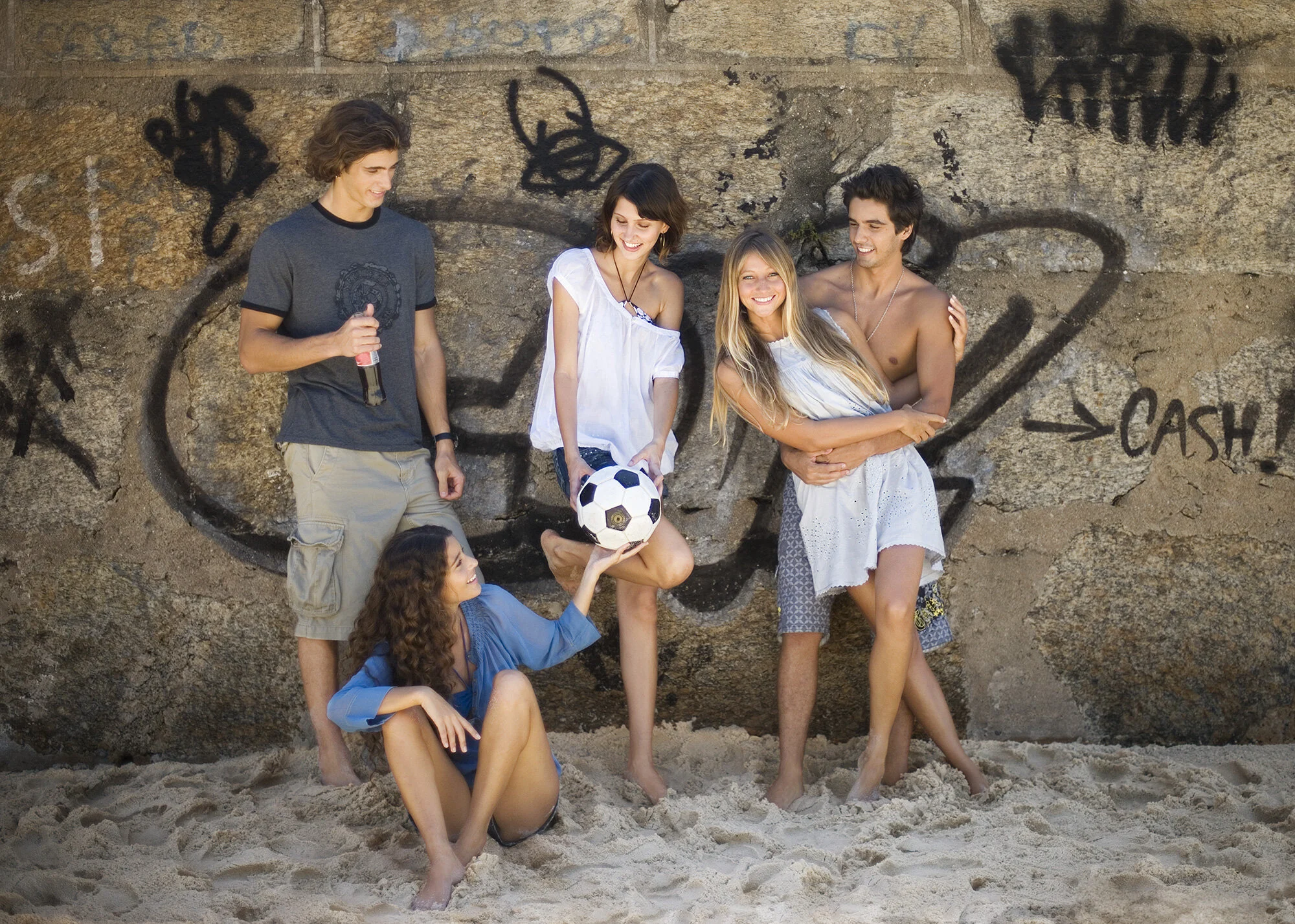 Group of five Brazilian friends on Ipanema beach, some holding drinks and a soccer ball, standing and sitting near graffiti on a stone wall, smiling and enjoying each other's company in Rio de Janeiro, Brazil.