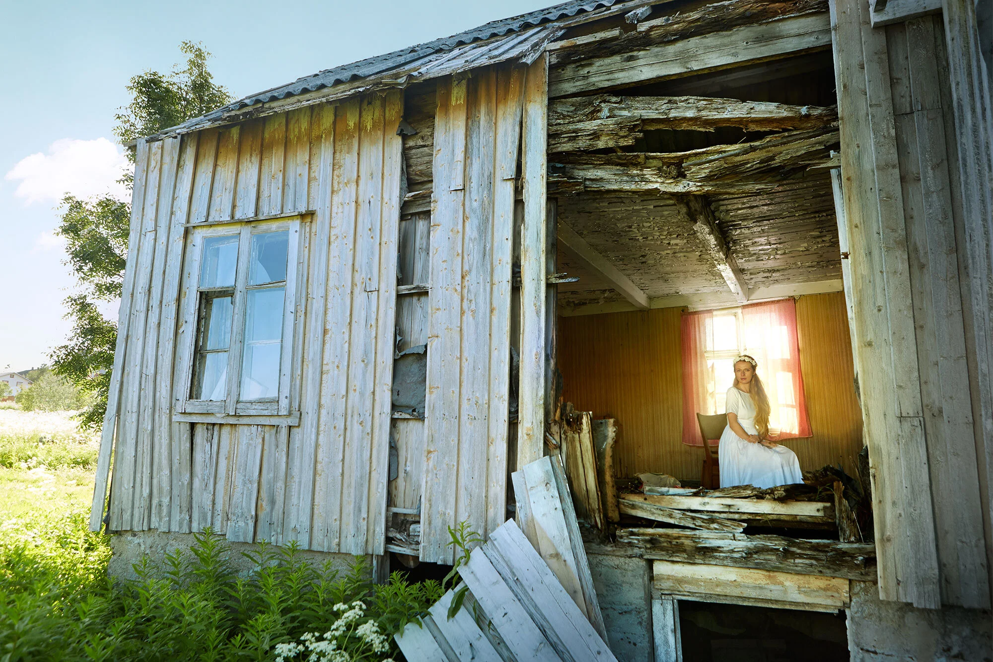 A young white woman with long hair in a white vintage dress sitting on a chair inside an old, damaged wooden house with broken walls and a window, illuminated by sunlight through a curtain in Lofoten, Norway.