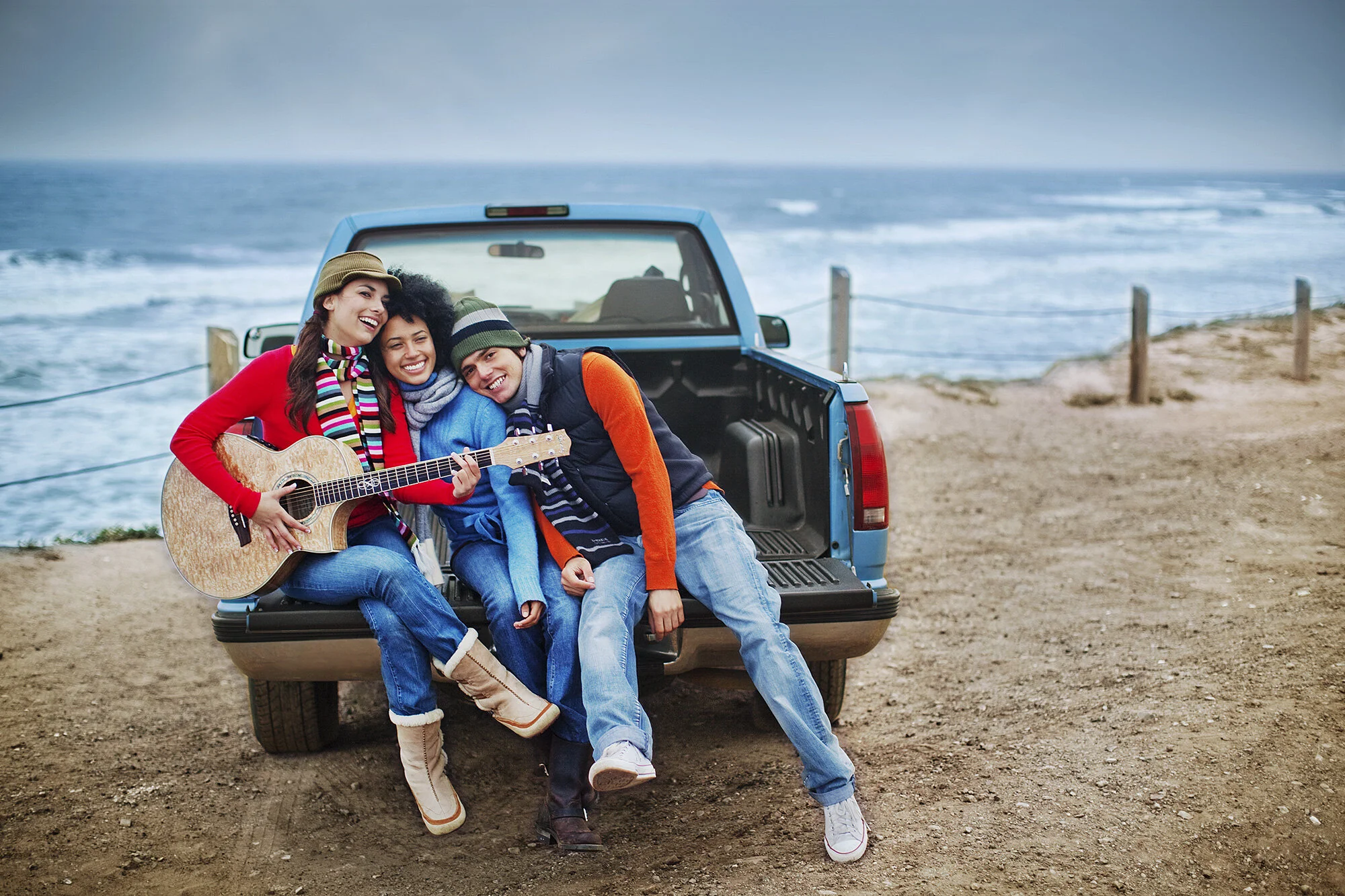 Three friends sitting on the back of a pickup truck on a beach in colorful wardrobe, smiling and enjoying each other's company. One person is playing an acoustic guitar. The background features the ocean and a cloudy sky at Ocean Beach in San Francis