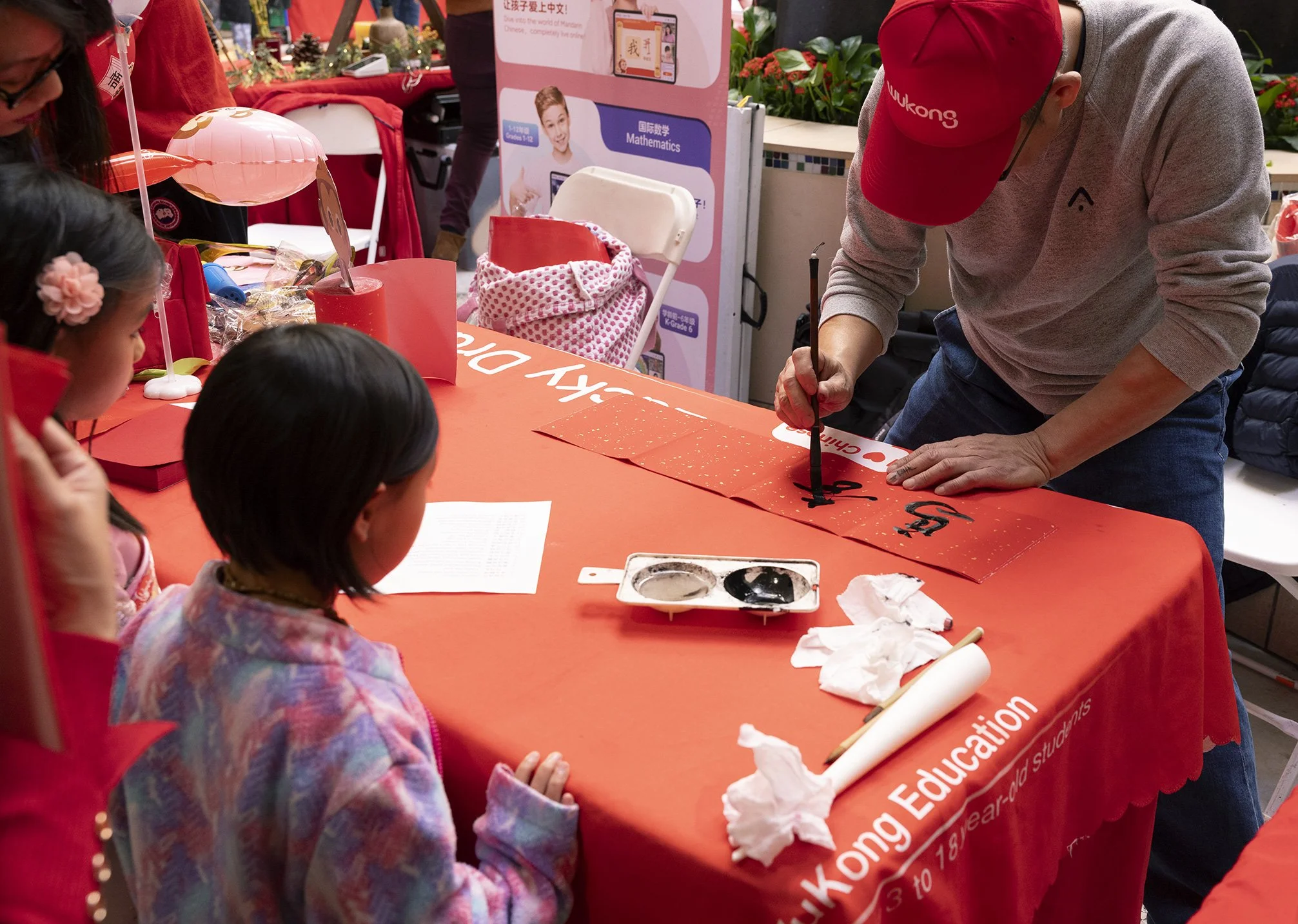 Man writing characters on a red piece of paper with young girls watching at Lunar Year celebration in San Francisco, CA.