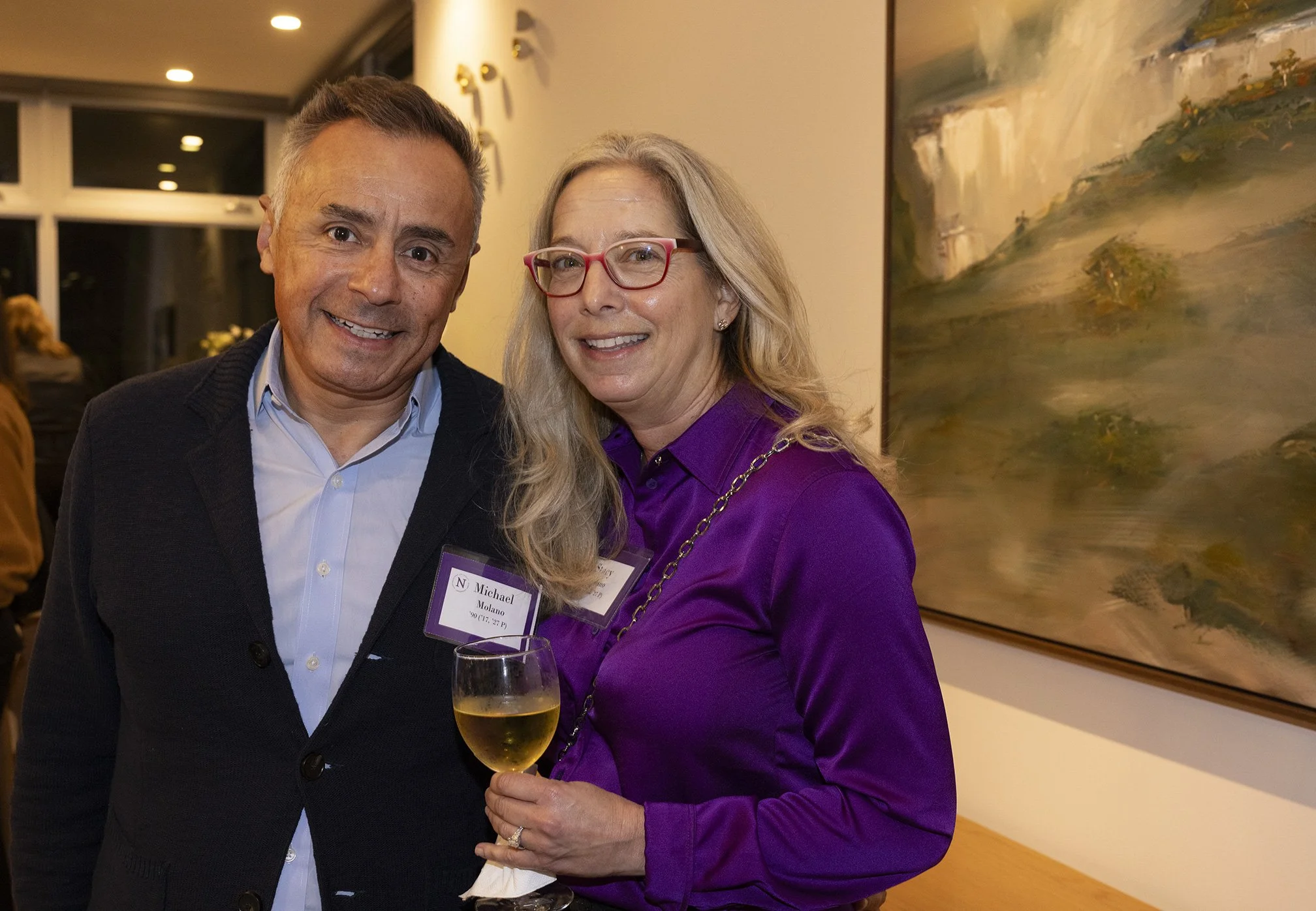 A couple standing together inside a house smiling at the camera. The woman is holding a glass of wine next to a large painting on the wall. Photographed for Northwestern University fundraiser.
