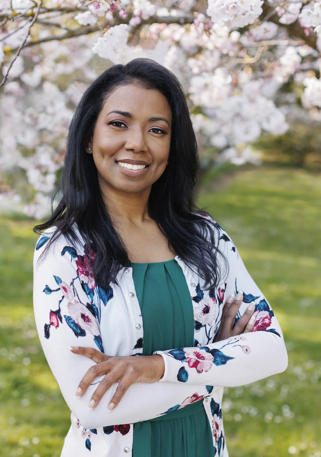 Young woman standing with her arms crossed wearing a green shirt and white floral cardigan, smiling at the camera with cherry blossoms in the background in the Botanical Garden in San Francisco, CA.