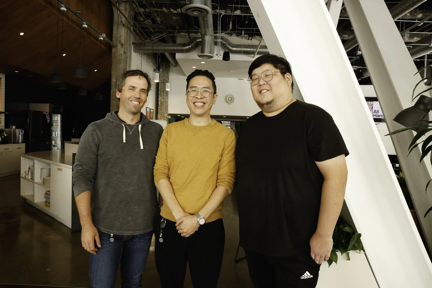 Three men smiling and standing close together indoors, in a modern space with exposed ductwork, a white partition, and a coffee station in the background at Brex Headquarter in San Francisco, CA.