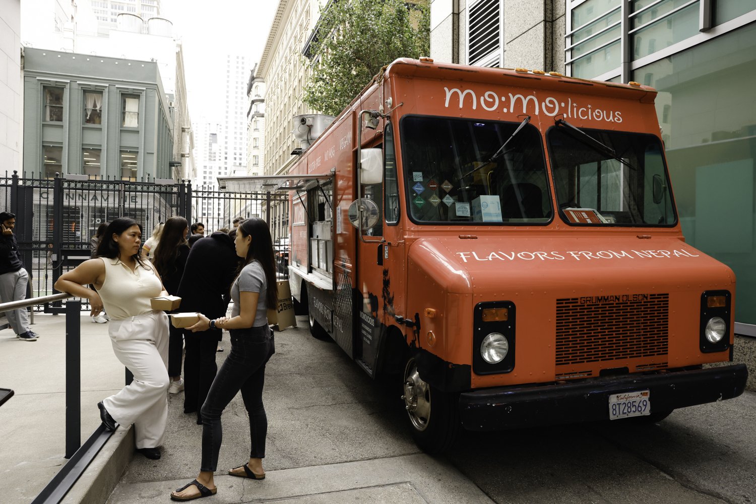 A red food truck with the words 'mo:mo:licious' and 'FLAVORS FROM NEPAL' parked on a city street, with people standing in line and interacting nearby Brex headquarter in San Francisco, CA.