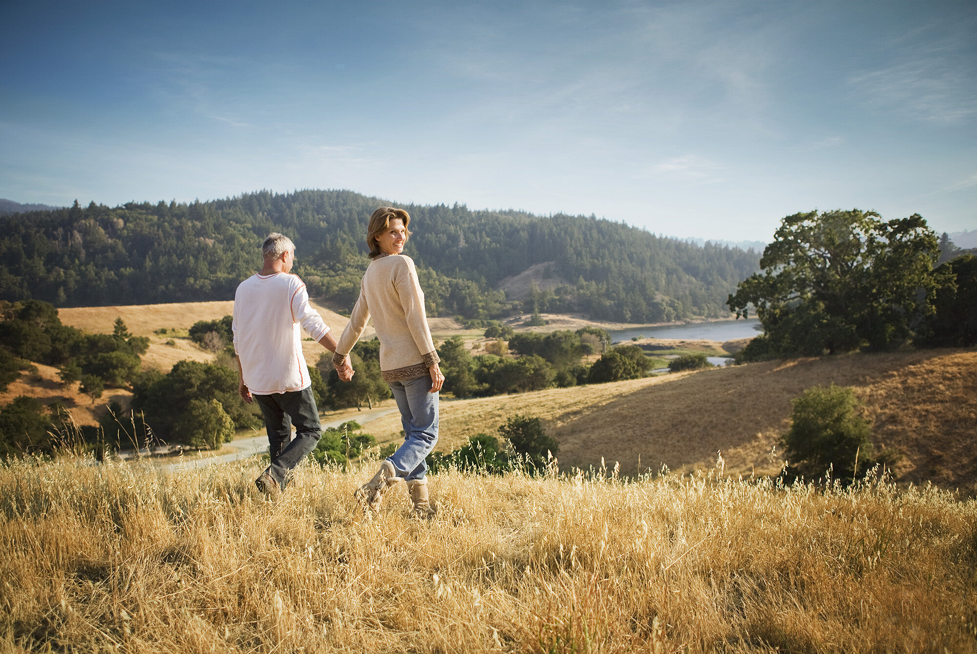 A white middle-aged couple holding hands and walking through a grassy field on a sunny day with a scenic landscape of hills, trees, and a lake in the background in San Francisco, CA.
