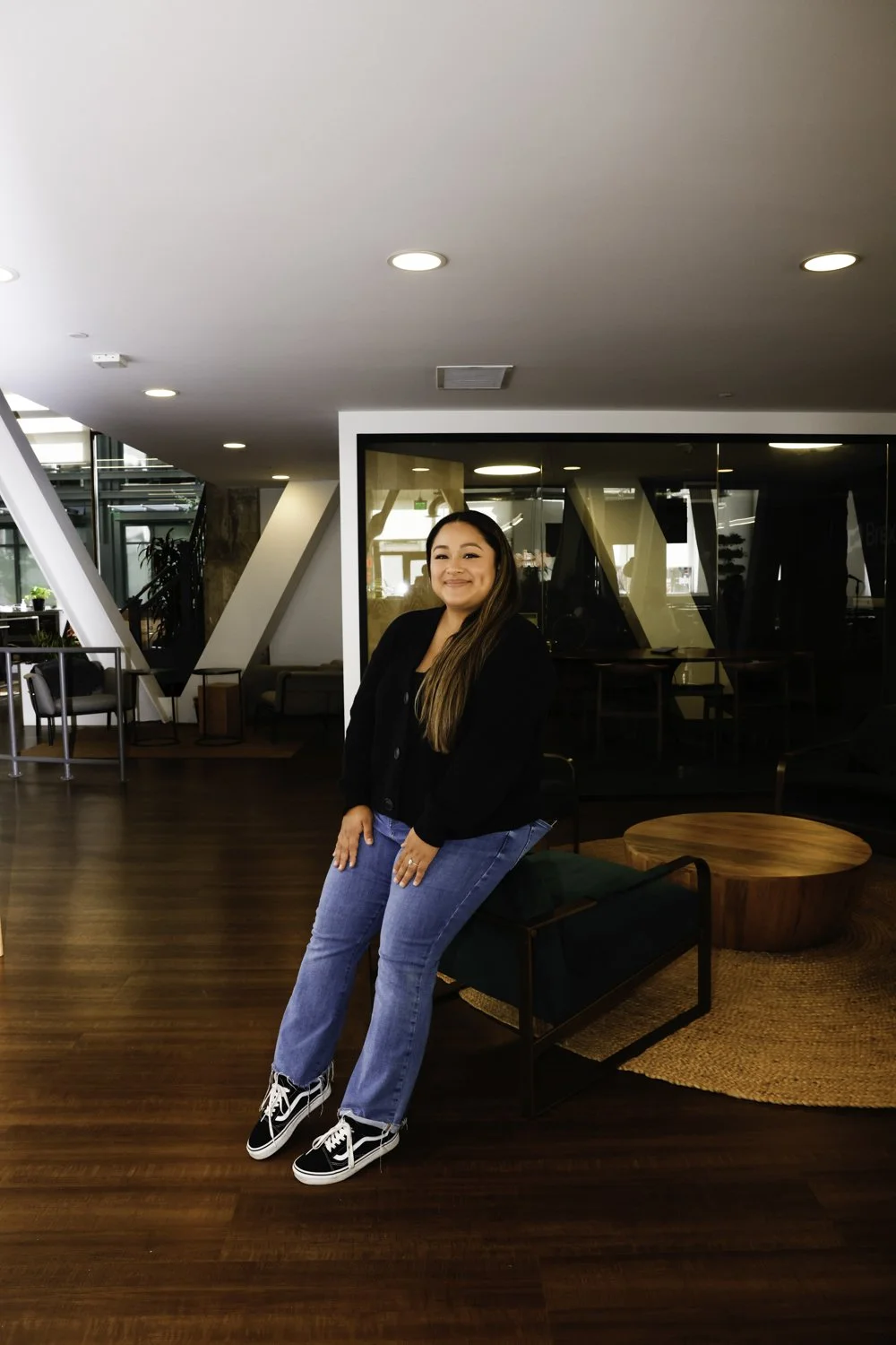 A woman with long brown hair, wearing a black jacket, blue jeans, and black and white sneakers, sitting on a green chair in a modern indoor space with wooden flooring and contemporary furniture at Brex Headquarter in San Francisco, CA.