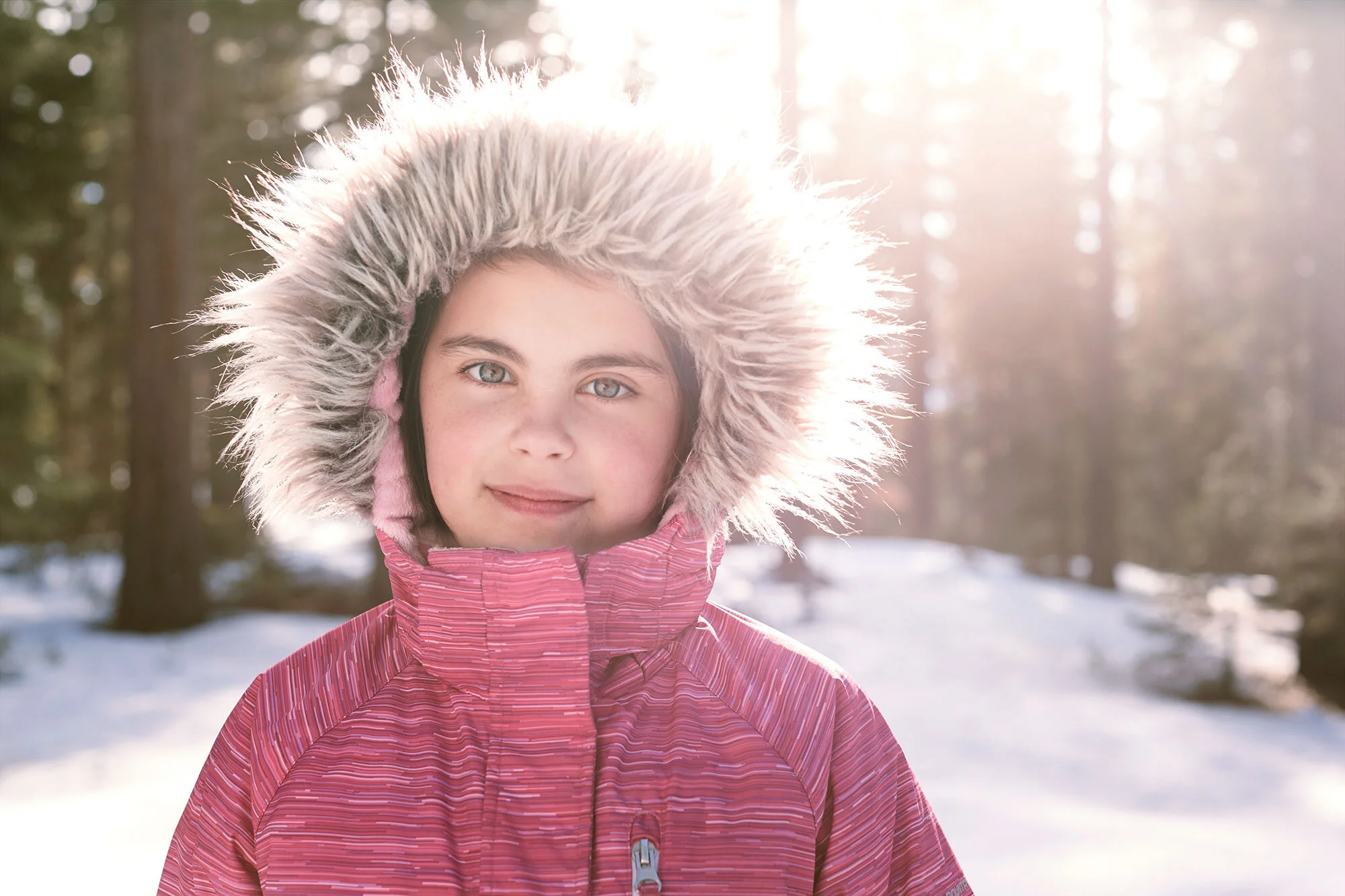 A girl in a pink winter coat with a fur-lined hood standing outdoors in a snow-covered forest, sunlight shining through the trees at Lake Tahoe, CA.
