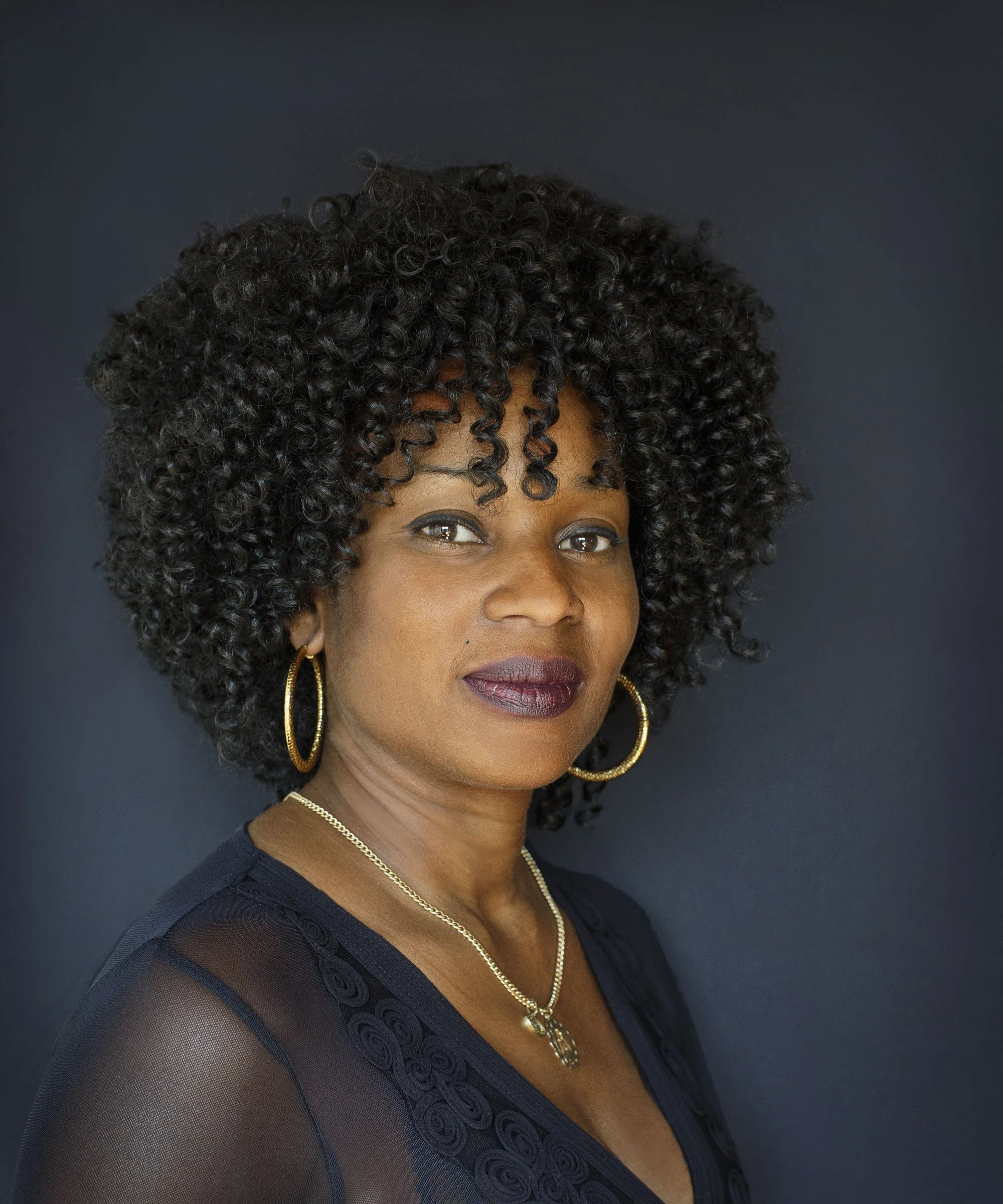 A close-up portrait of a Black woman, short curly black hair, wearing gold hoop earrings, a gold necklace, and a black sheer top with embroidered swirling patterns, against a dark background in Norway