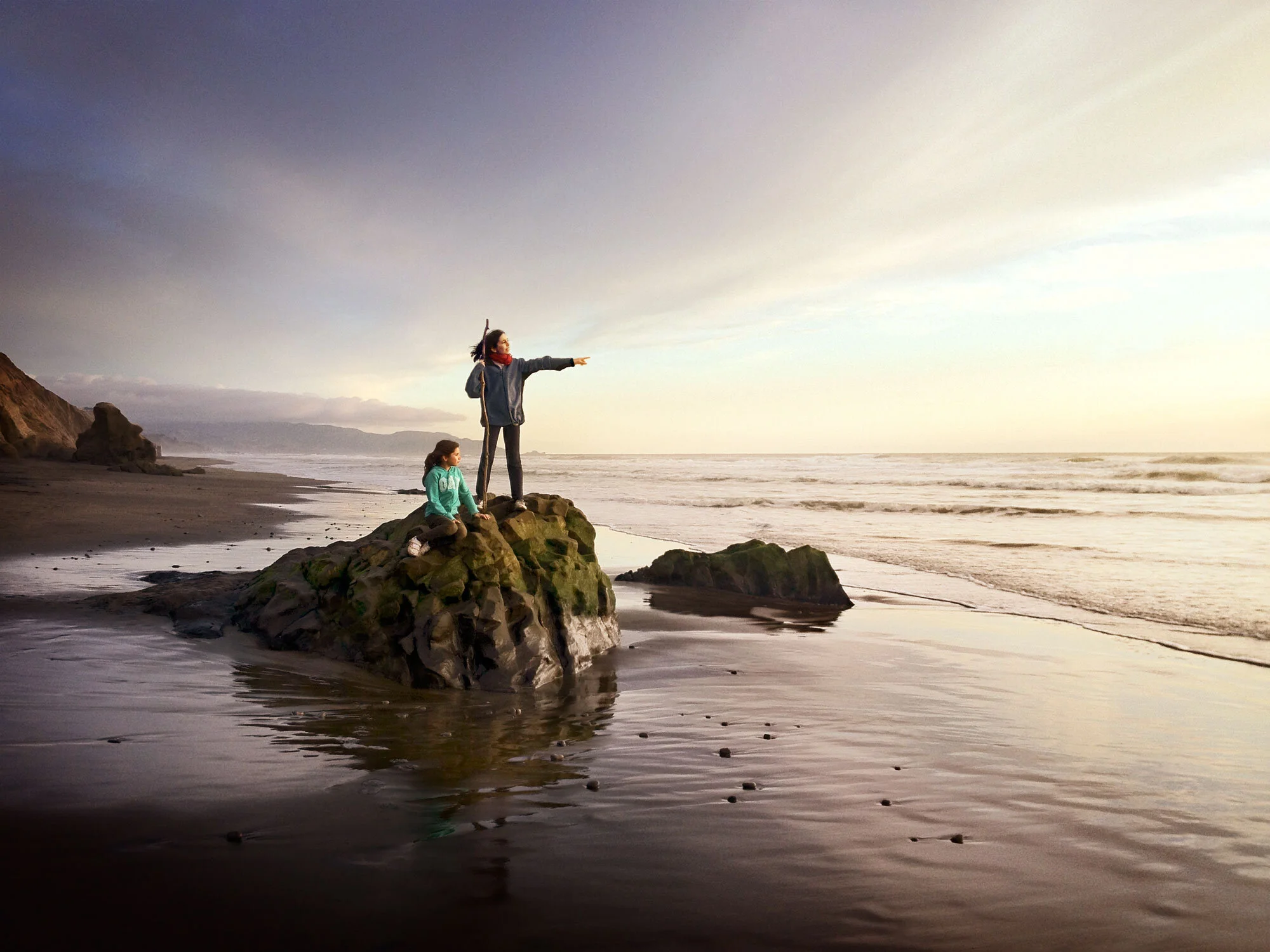 Two young girls one standing and one sitting on a rock on the beach at Fort Funston in San Francisco, CA at sunset with purple and yellow light.