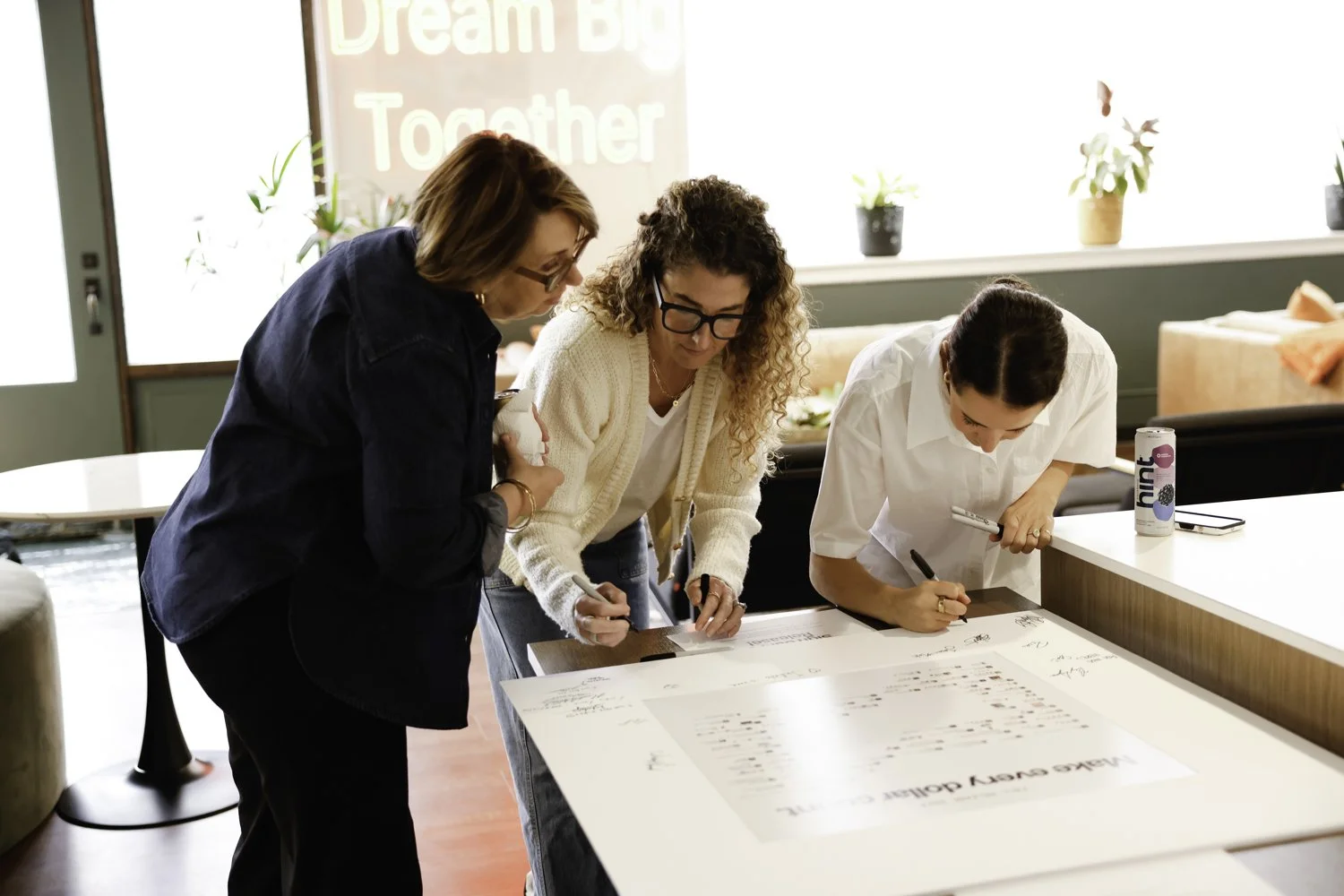 Three women gather around a large table, writing or signing documents at Brex Headquarter in San Francisco, CA.