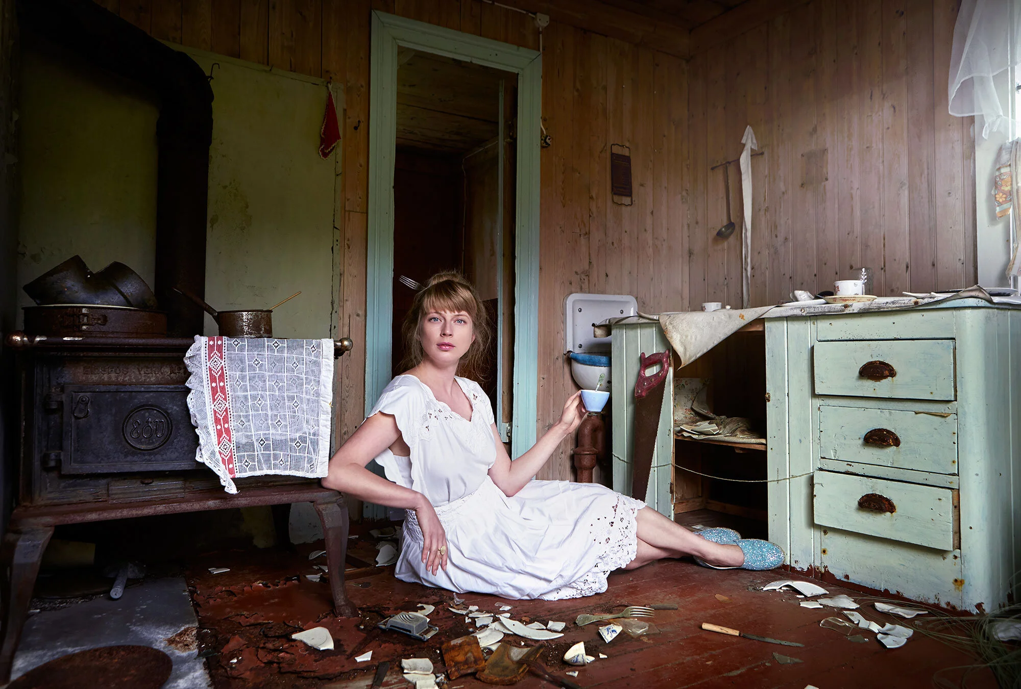 Woman with her hair in a bun with a fork through it with a white vintage dress sitting on the floor of a cluttered, old kitchen, holding a small cup, with broken dishes scattered around, in front of an old stove and a green cabinet at Lofoten, Norway