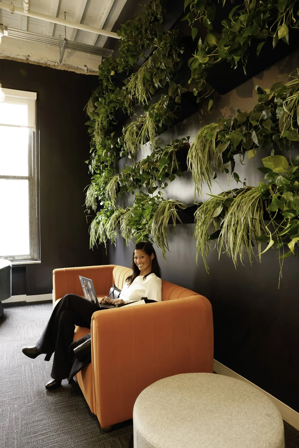 A woman smiling and working on her laptop while sitting on an orange sofa in a room with dark walls, large green hanging plants, a window letting in natural light, and a beige round ottoman in the foreground Brex headquarter in San Francisco, CA.