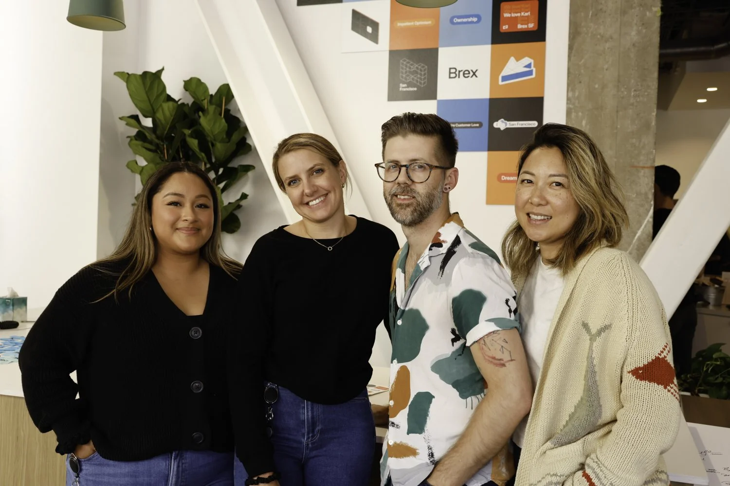 Four diverse people standing together inside an office, smiling, with a large colorful poster and plants in the background Brex headquarter in San Francisco, CA.